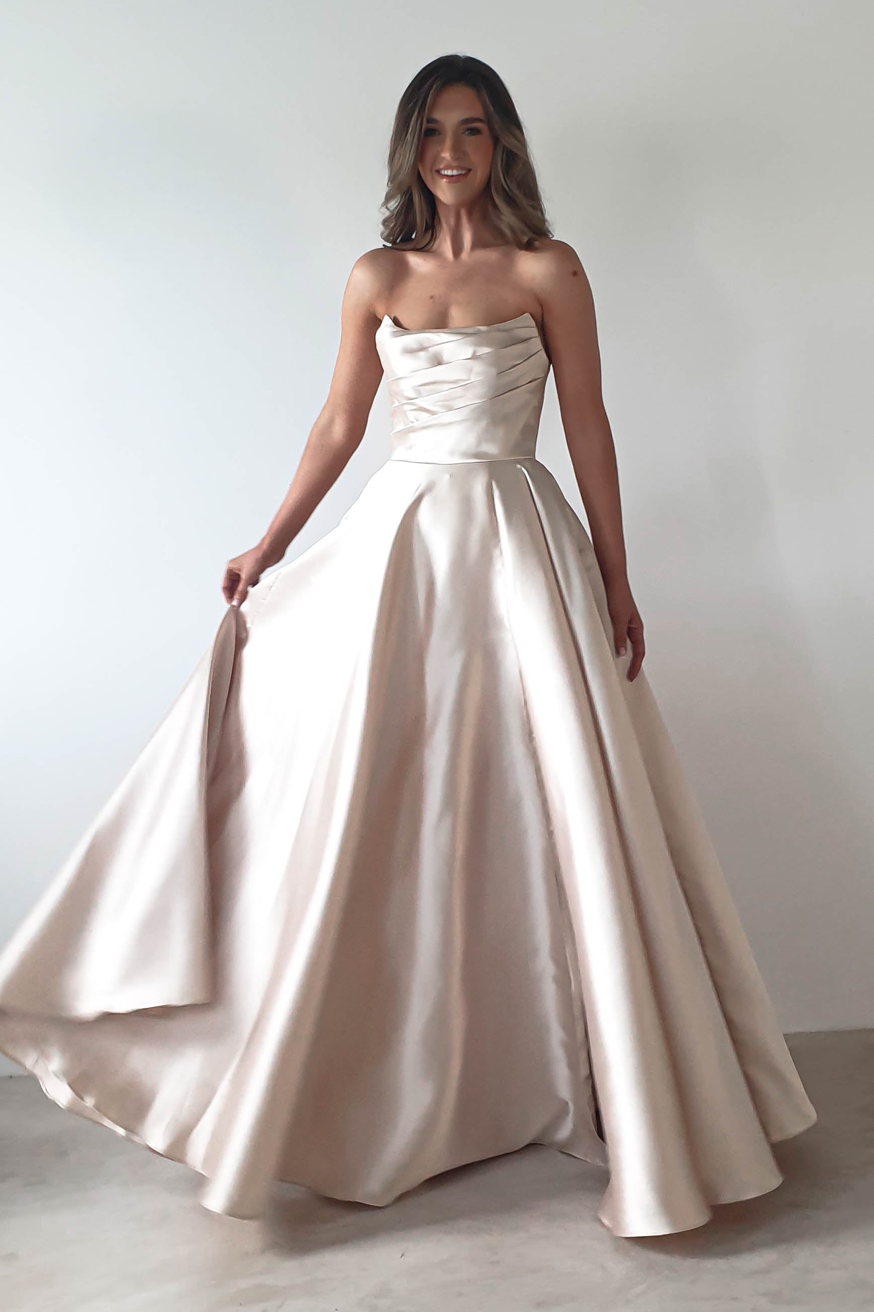 At a formal event, a woman graces the scene in an elegant, strapless cream-colored gown. The dress boasts a fitted bodice with ruching and an A-line skirt. She smiles warmly, gently holding the sides of her voluminous attire against a plain backdrop.
