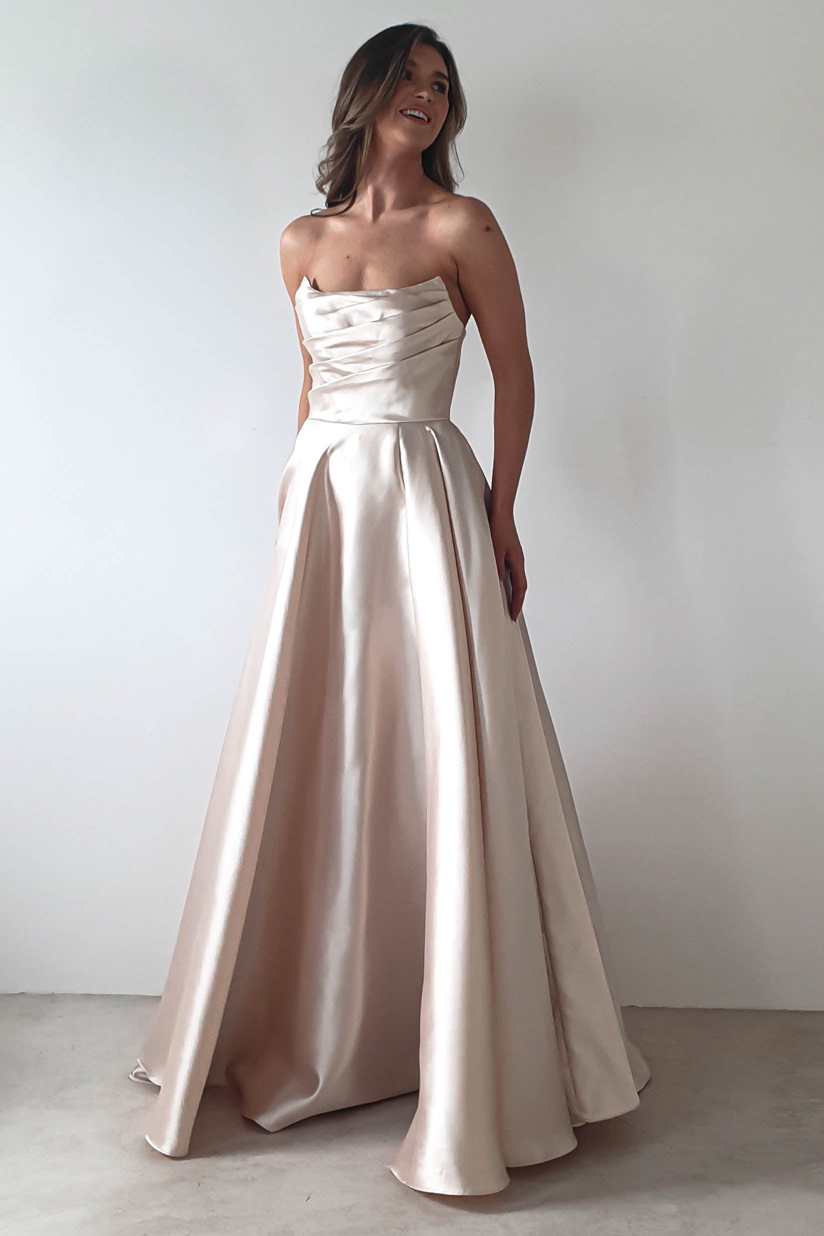 A person wearing a strapless, elegant satin gown poses gracefully against a plain white background. The A-line skirt adds volume to the outfit, perfect for a formal event. With long hair cascaded down their shoulders, they appear to be smiling warmly.