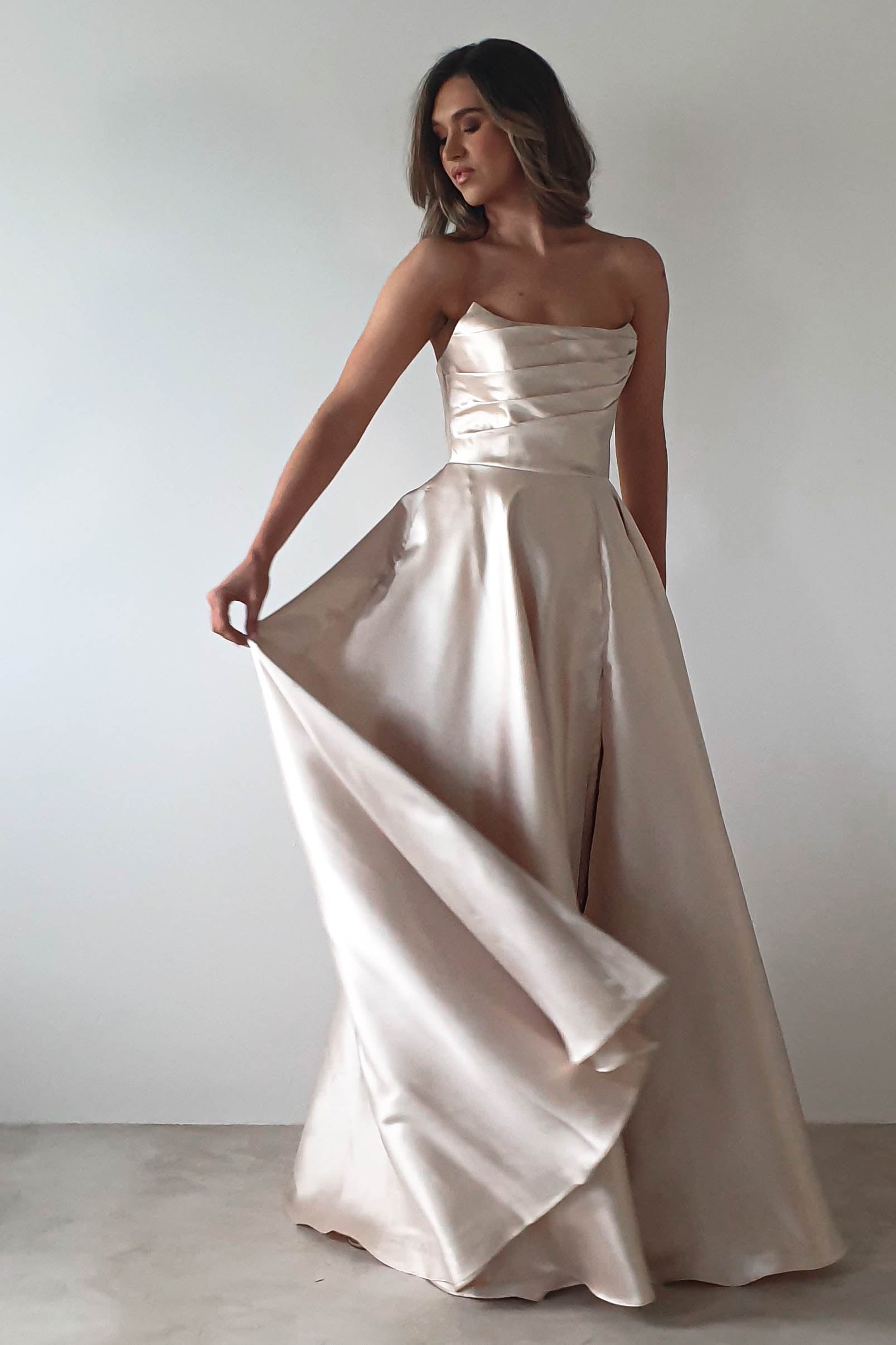 A woman in a strapless, flowing champagne-colored gown poses elegantly against a plain white background, perfect for a formal event. The dress features a fitted bodice and an A-line skirt, with her hand holding one side to create graceful movement.