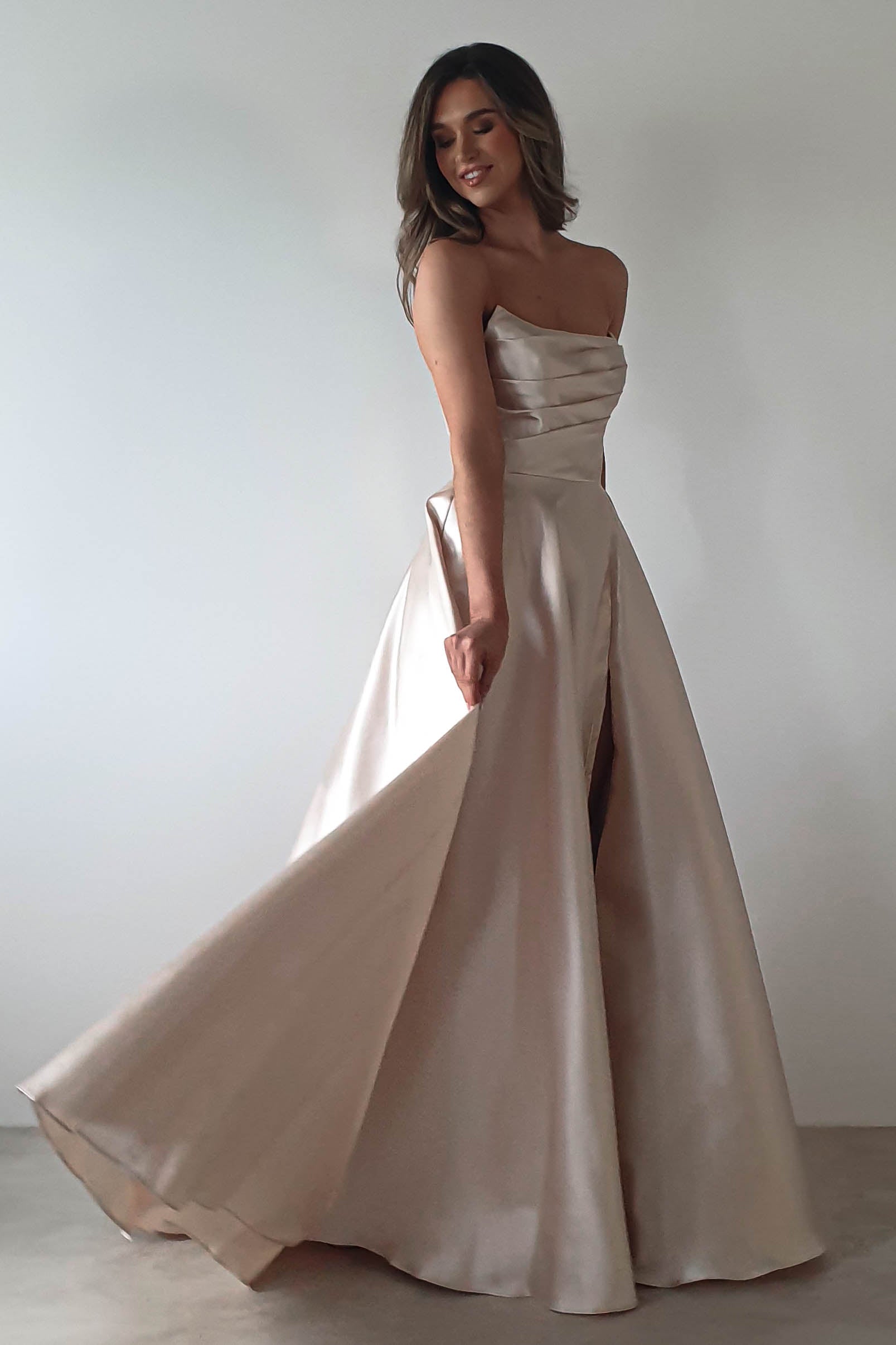 A woman in a strapless, flowing beige gown poses gracefully against a plain background, ready for a formal event. The dress has an A-line skirt with a subtle sheen. She has long hair and smiles softly while turning slightly to the side.