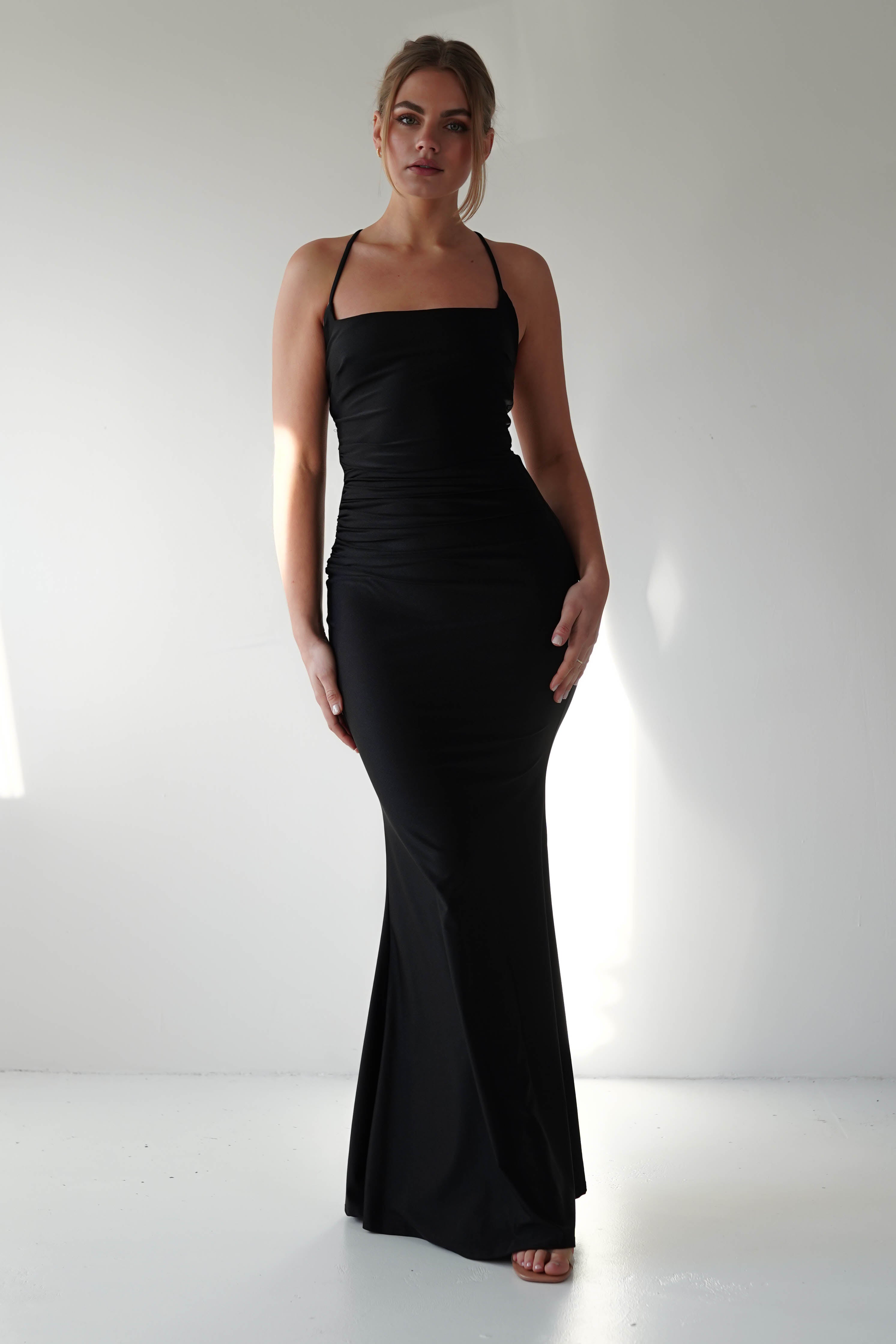 A woman stands against a plain white background wearing a fitted black gown with thin straps and a mermaid-style skirt. She looks toward the camera with a neutral expression, her hair pulled back. Light and shadows are visible on the wall behind her.