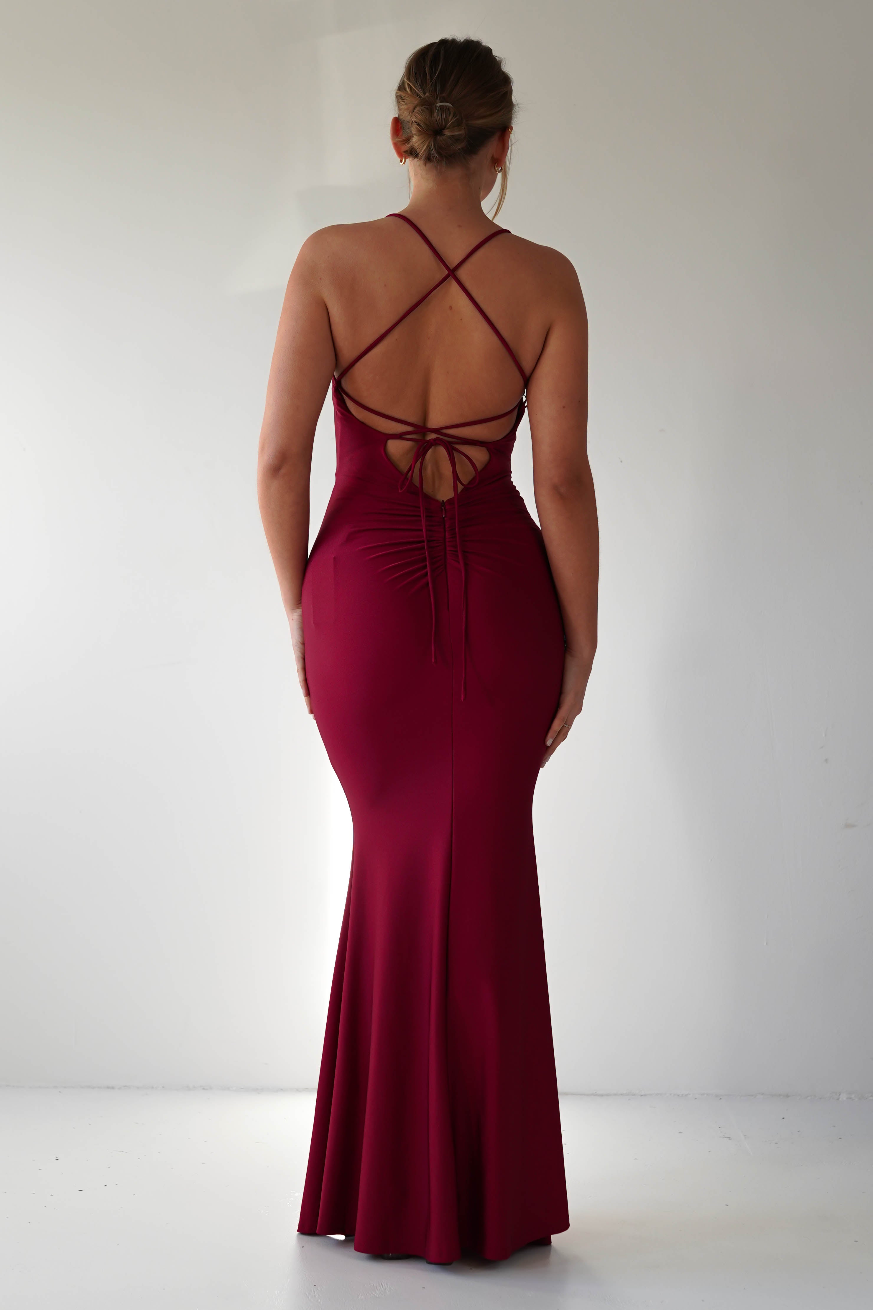 A woman stands facing away, wearing a floor-length, mermaid-style deep red gown with crisscross straps and an open back. Her hair is styled in a low bun as she poses against a plain white background, perfect for formal occasions.