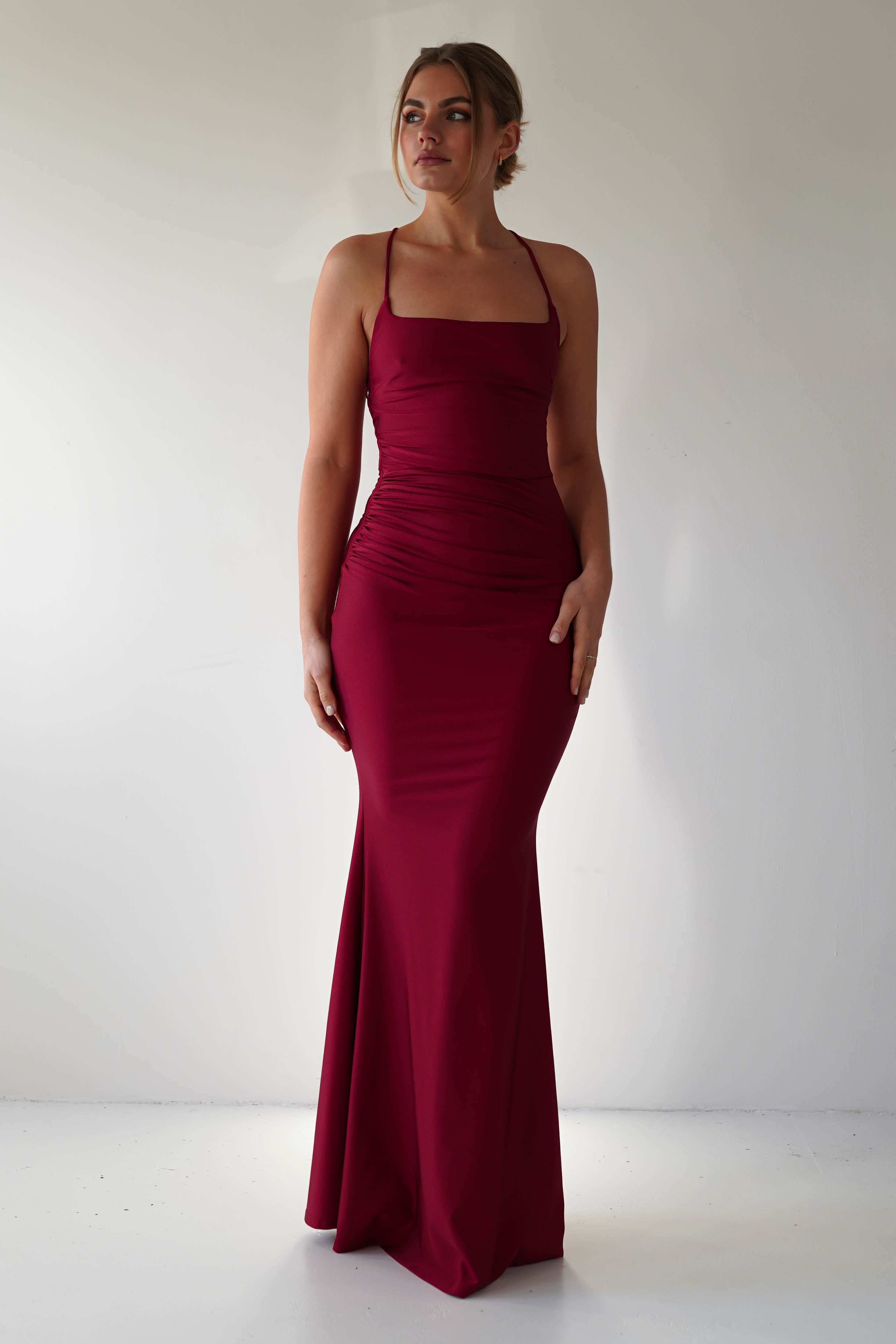 A woman stands against a plain light-colored background wearing a sleeveless, mermaid-style, floor-length burgundy gown with thin straps. She looks slightly to the side with one hand resting on her hip—a perfect look for formal occasions.