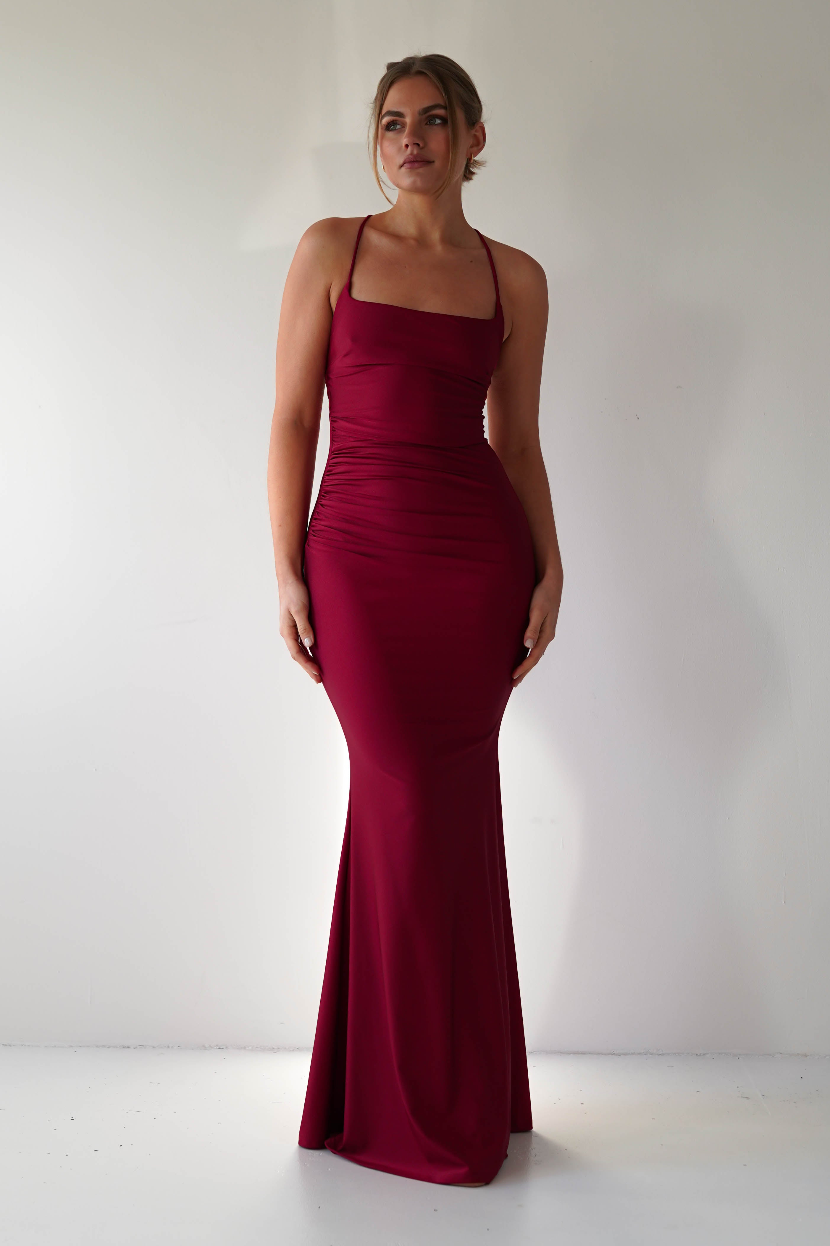 A woman stands against a plain white background in a fitted, sleeveless maroon mermaid-style gown with thin straps and a floor-length hem—perfect for formal occasions. Her hair is styled up as she looks slightly to the side.