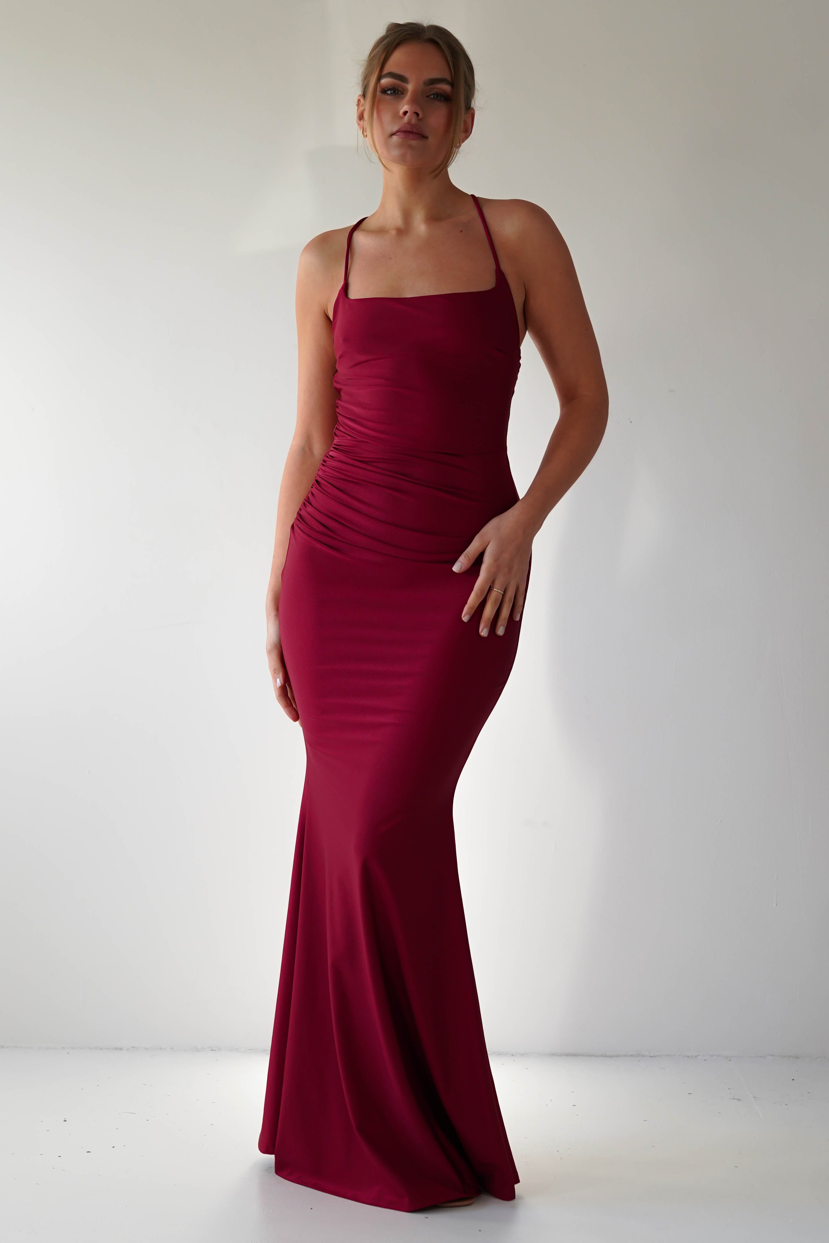 A person stands against a plain white background wearing a fitted, floor-length mermaid-style burgundy gown with thin straps and subtle ruching on one side, posing with one hand on their hip—perfect for formal occasions.