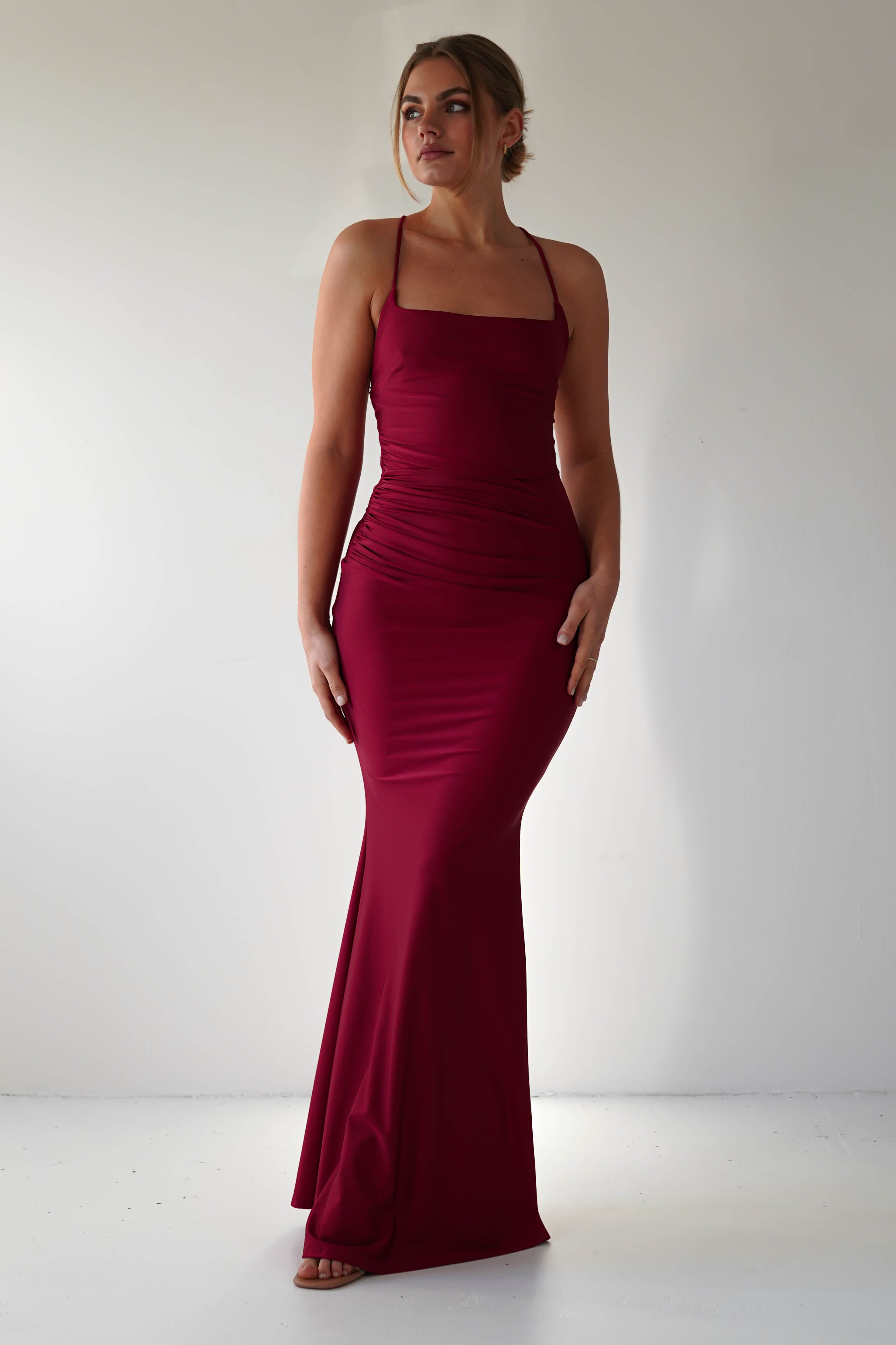 A woman wearing a fitted, floor-length mermaid-style burgundy gown stands against a plain light background, her hair styled up and her arms resting by her sides—perfect for formal occasions.