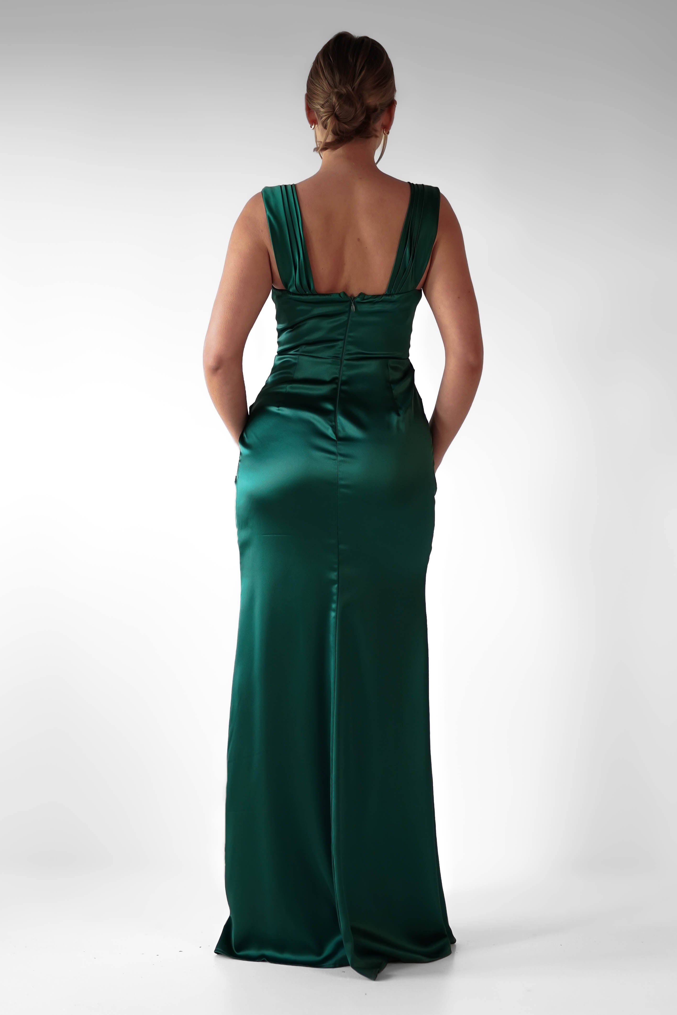 A woman stands facing away, wearing a formal evening gown in emerald green satin with flattering ruching and a deep V-back. Her hair is styled in a low updo, and she stands against a plain white background.