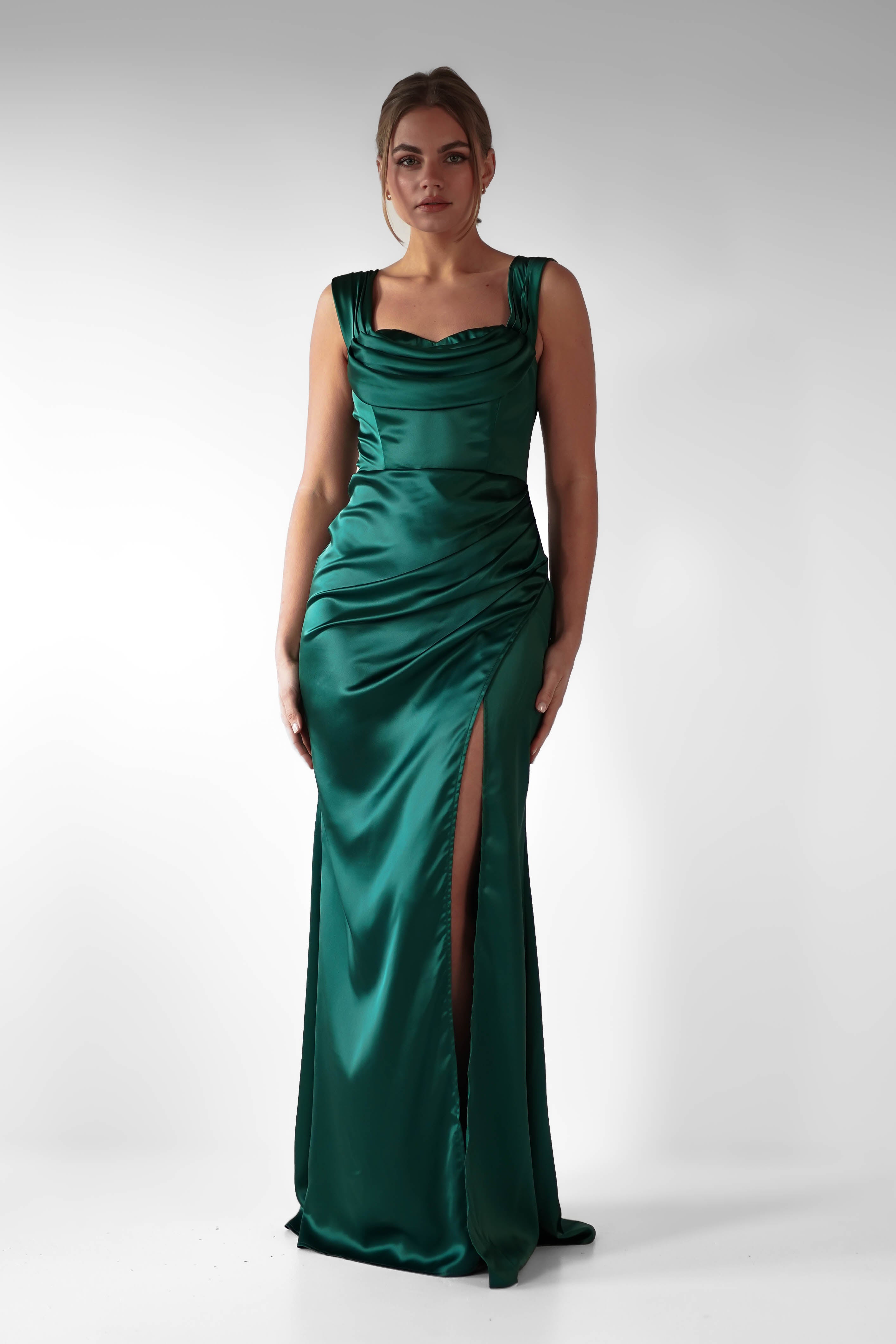 A woman stands against a plain background wearing a formal evening gown in emerald green satin, featuring flattering ruching at the draped bodice and a high thigh slit. She has her hair styled back and looks directly at the camera.