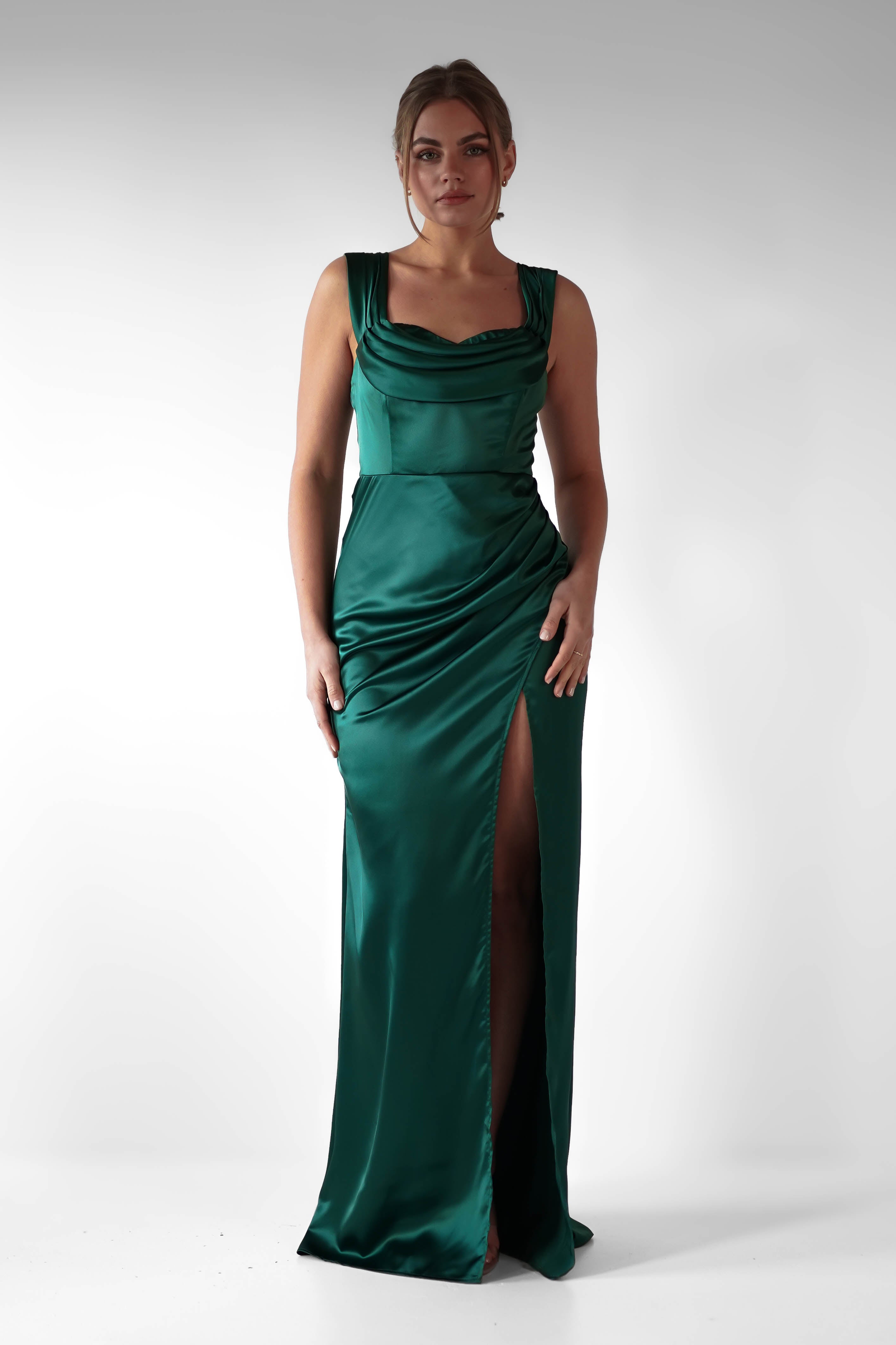 A woman stands against a plain white background wearing a sleeveless, floor-length emerald green formal evening gown with flattering ruching and a high slit on one side.