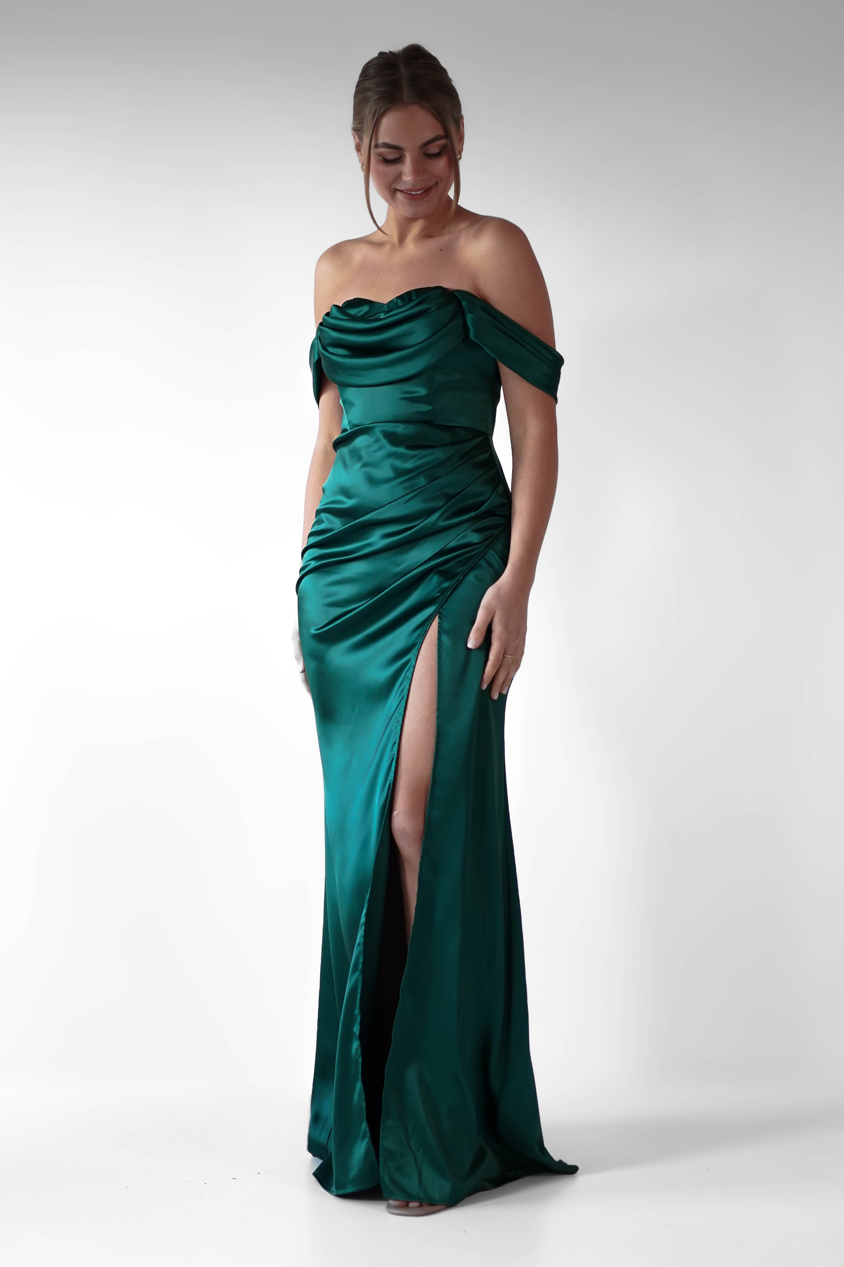 A woman stands in a studio wearing a formal evening gown—an emerald green, off-the-shoulder dress with a high slit and flattering ruching. She smiles, looking down, her hair styled in a loose updo against a plain white background.