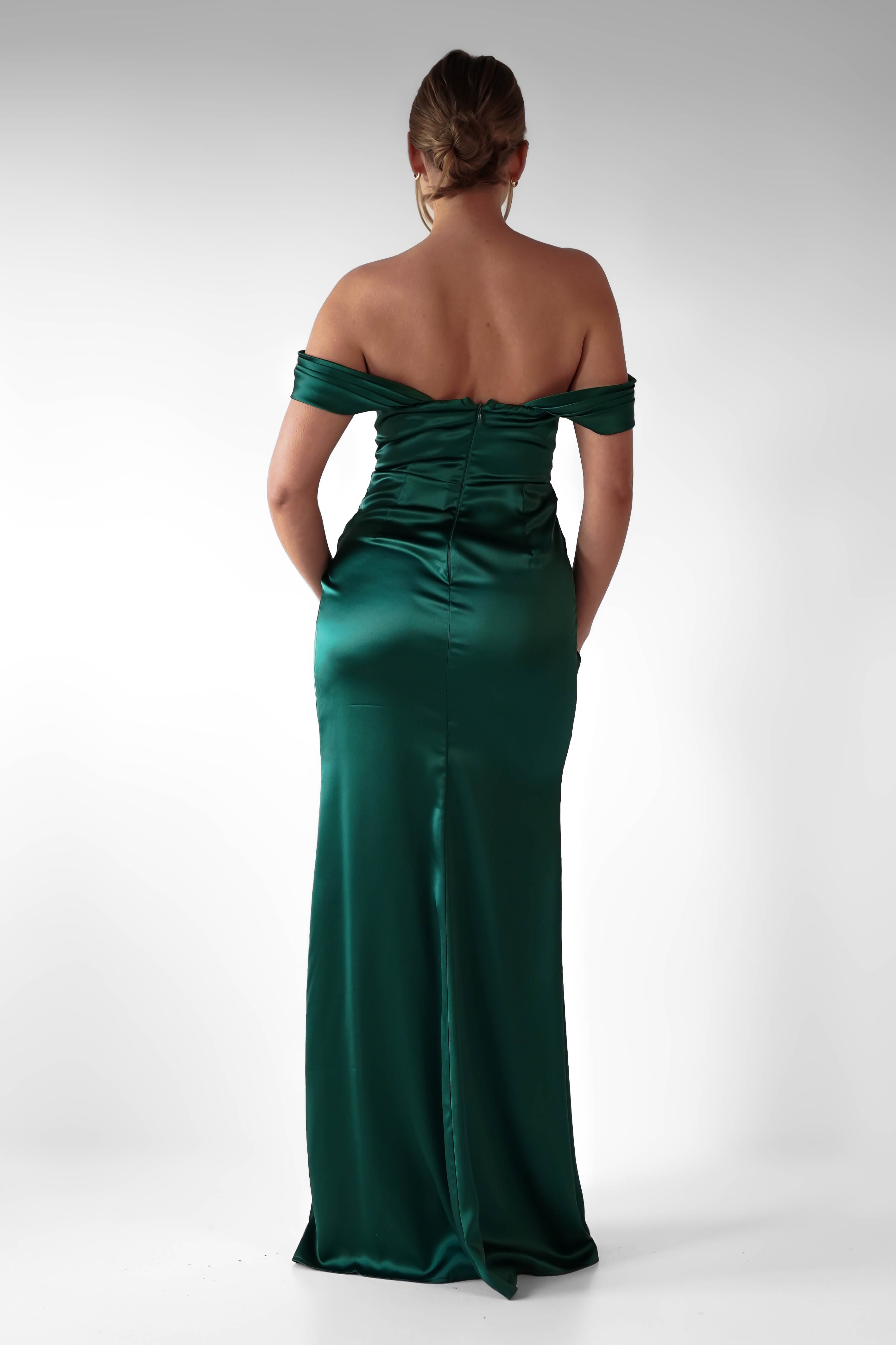 A woman stands with her back to the camera, wearing an off-the-shoulder formal evening gown in emerald green satin. Her hair is styled in an updo, and her hands rest casually in the flattering ruching of the dress’s pockets.