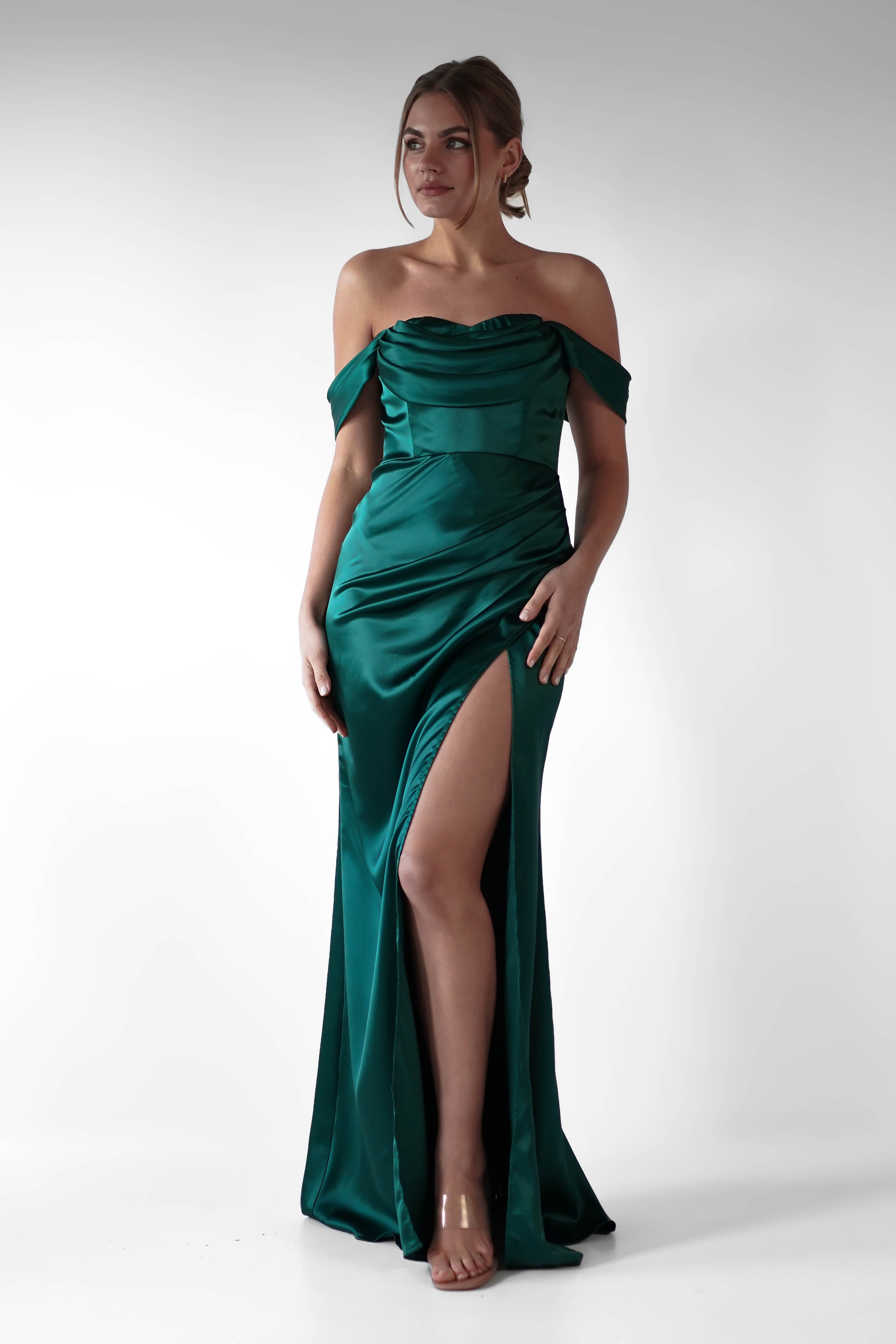 A woman wearing an off-the-shoulder dress in emerald green satin, featuring flattering ruching on the bodice and a high slit, stands barefoot against a plain white background.
