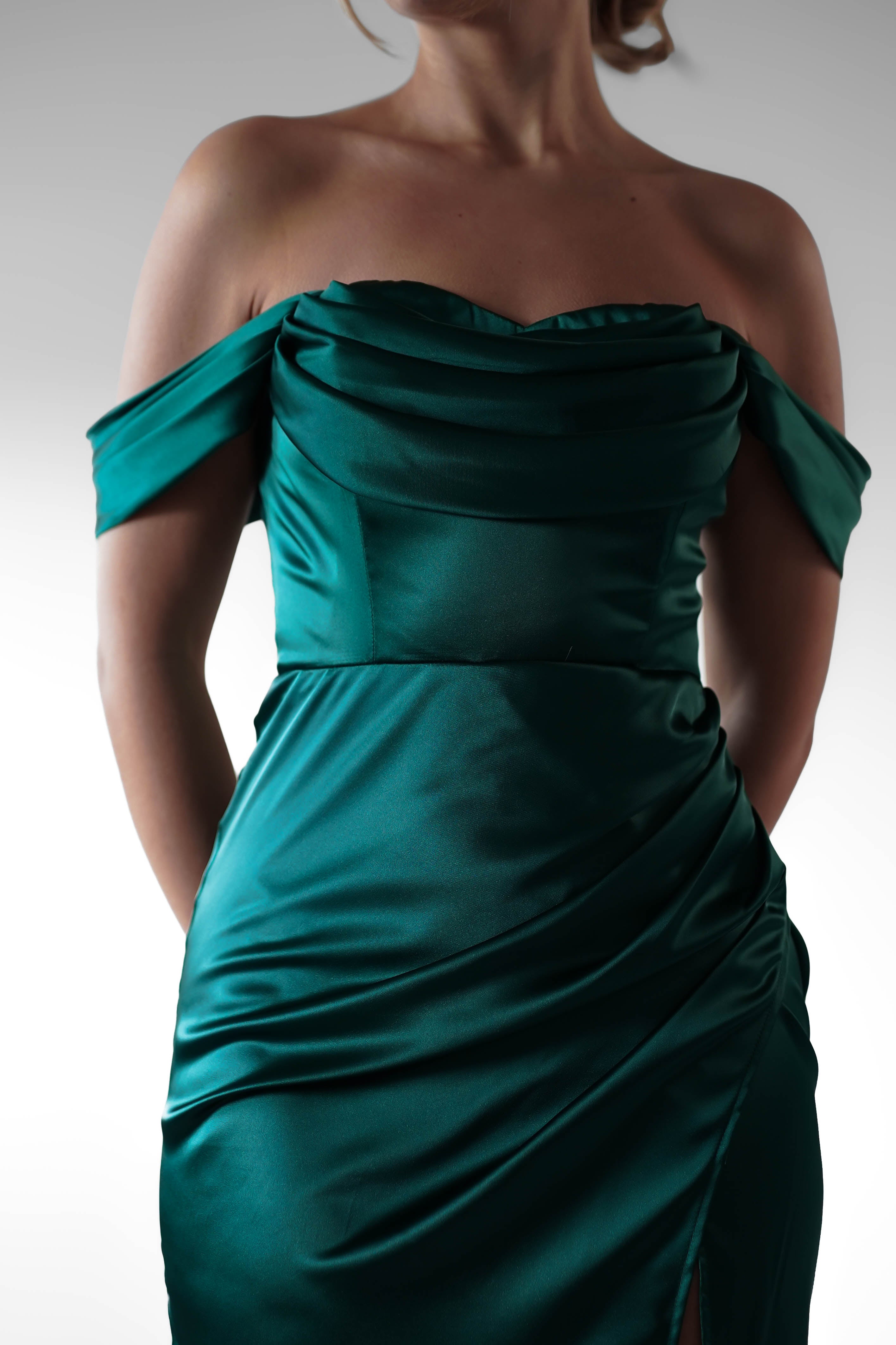A woman in an off-the-shoulder, deep teal satin formal evening gown with flattering ruching stands with her hands behind her back against a plain background. Her head is partially out of frame.