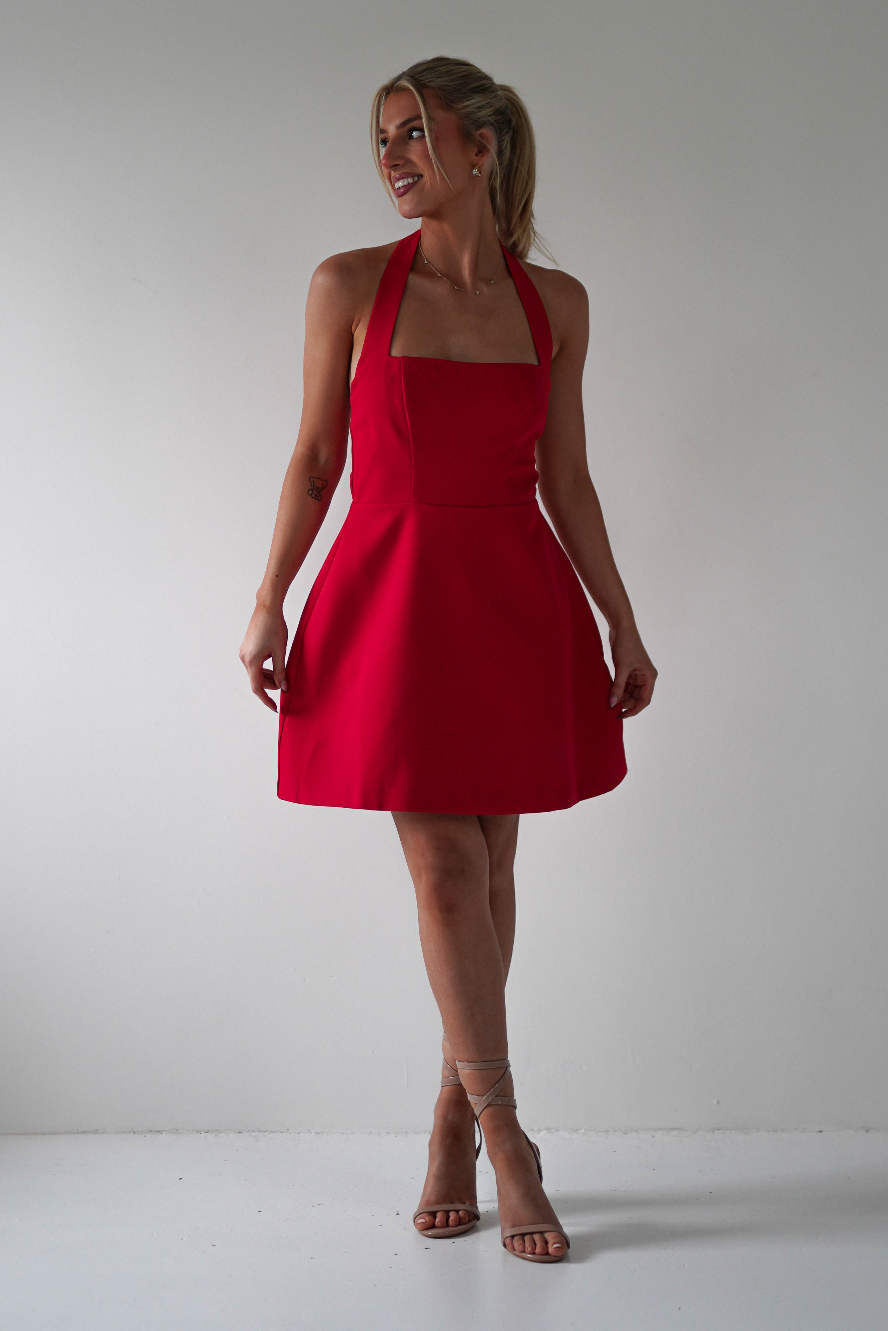 A person in a red halter dress smiles and poses against a white background, reminiscent of a Christmas party dress. They are wearing strappy beige high heels and have their hair tied back in a ponytail.