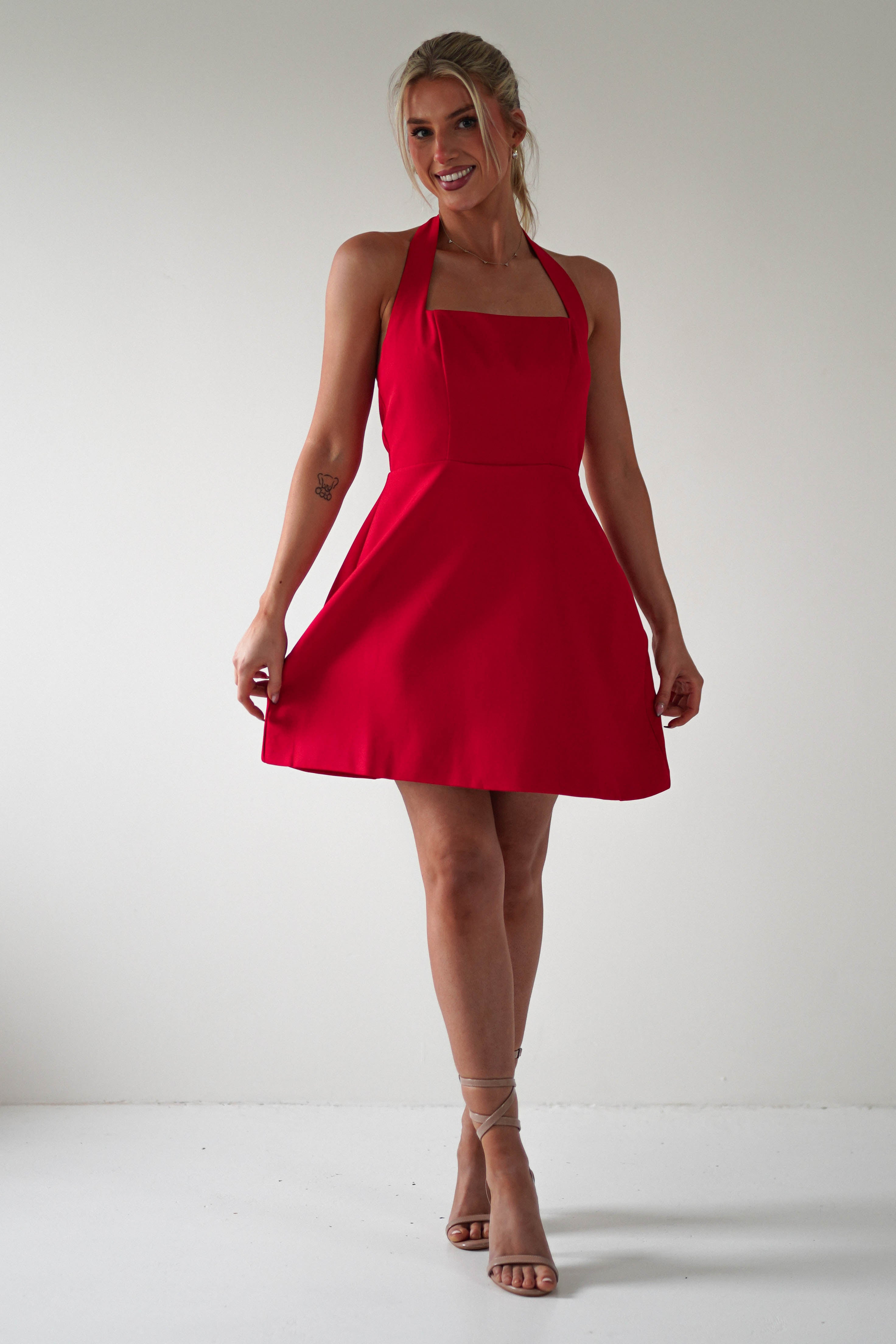 A person wearing a sleeveless red wedding guest dress and beige high-heeled sandals stands against a plain white background. They appear to be smiling and holding the sides of the dress.