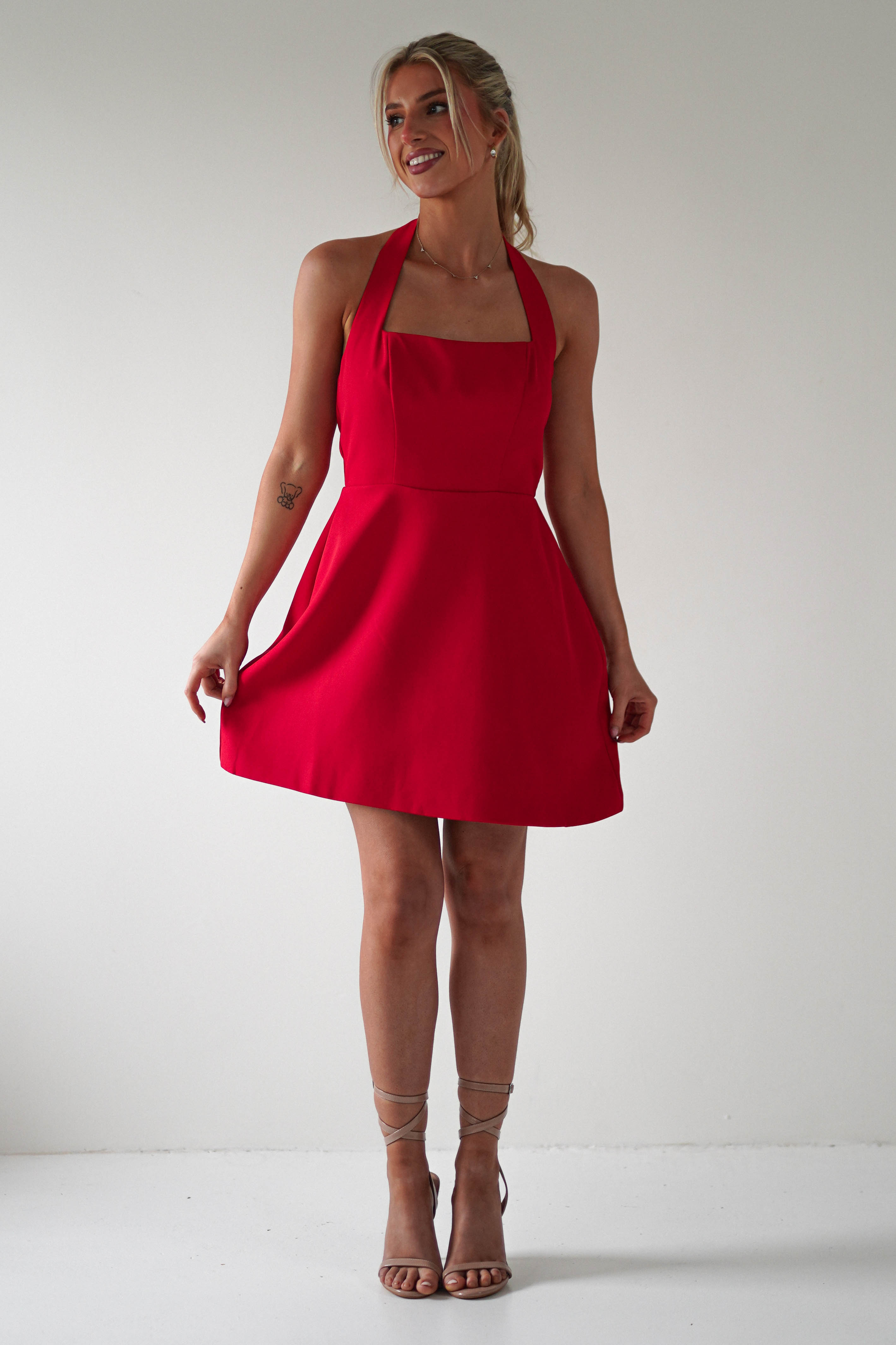 A woman stands smiling in a sleeveless red dress with a halter neck, perfect for a Christmas party. She holds the sides of her dress, wearing strappy high heels with her hair styled in an elegant updo. The plain white background enhances her festive elegance.