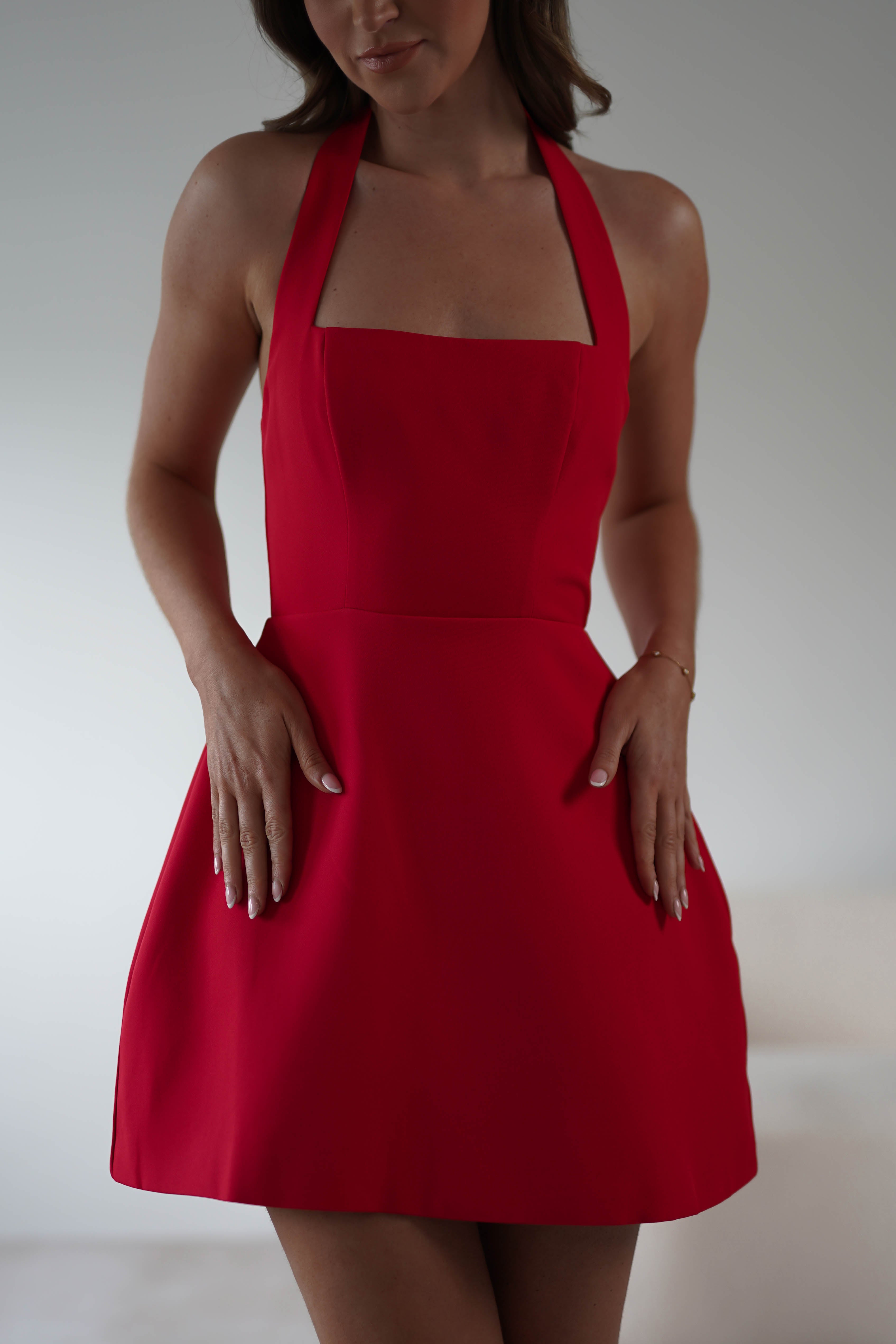 A person elegantly dons a sleeveless red dress with a square neckline and halter straps, perfect as a Christmas party dress. Their hands rest gently on the sides of the dress, while the background remains artfully out of focus.