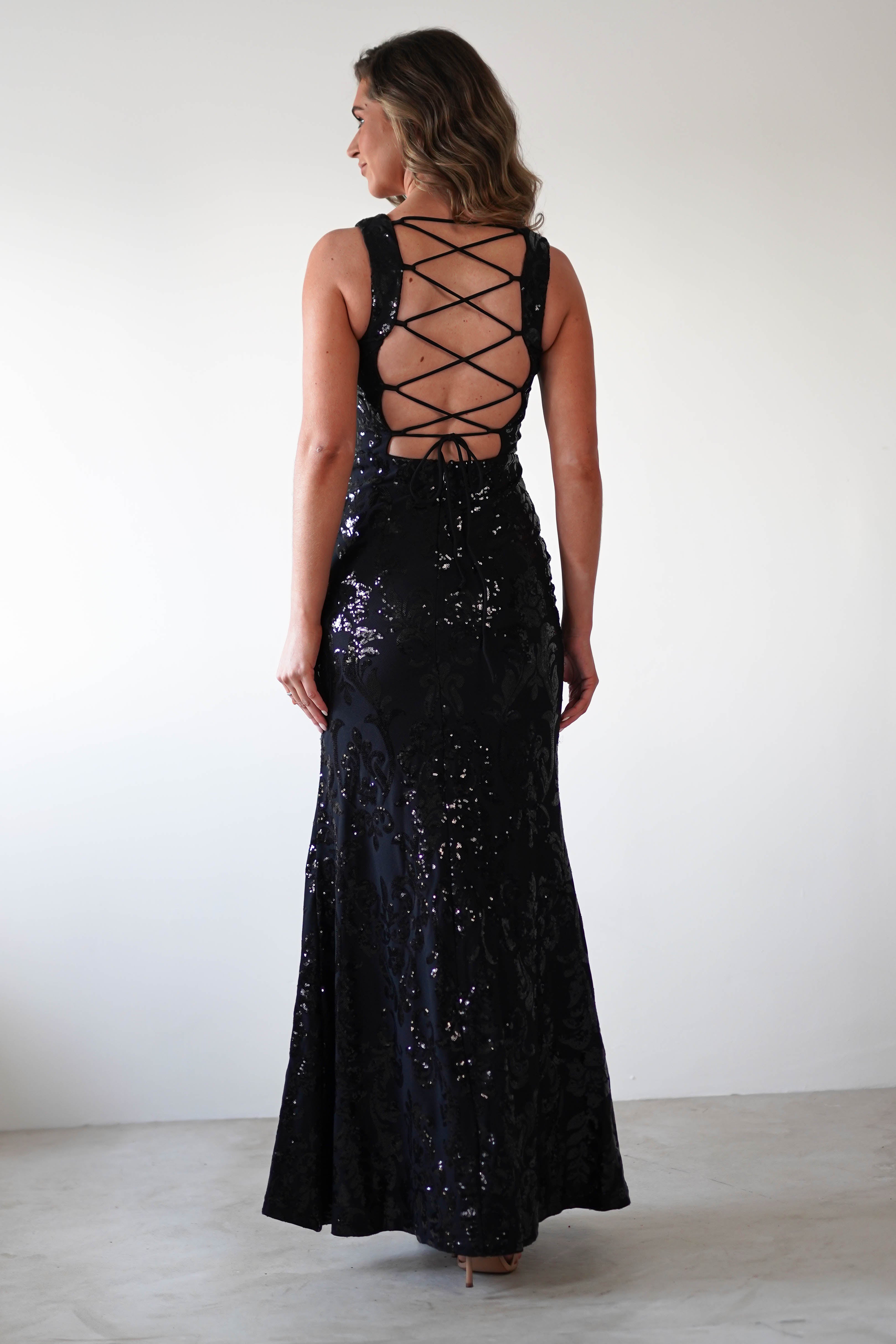A woman in a fitted black gown stands with her back to the camera. The square neckline of the dress complements its lace-up design and shimmering sequin detailing. Her wavy hair cascades elegantly as she poses against a plain white background.