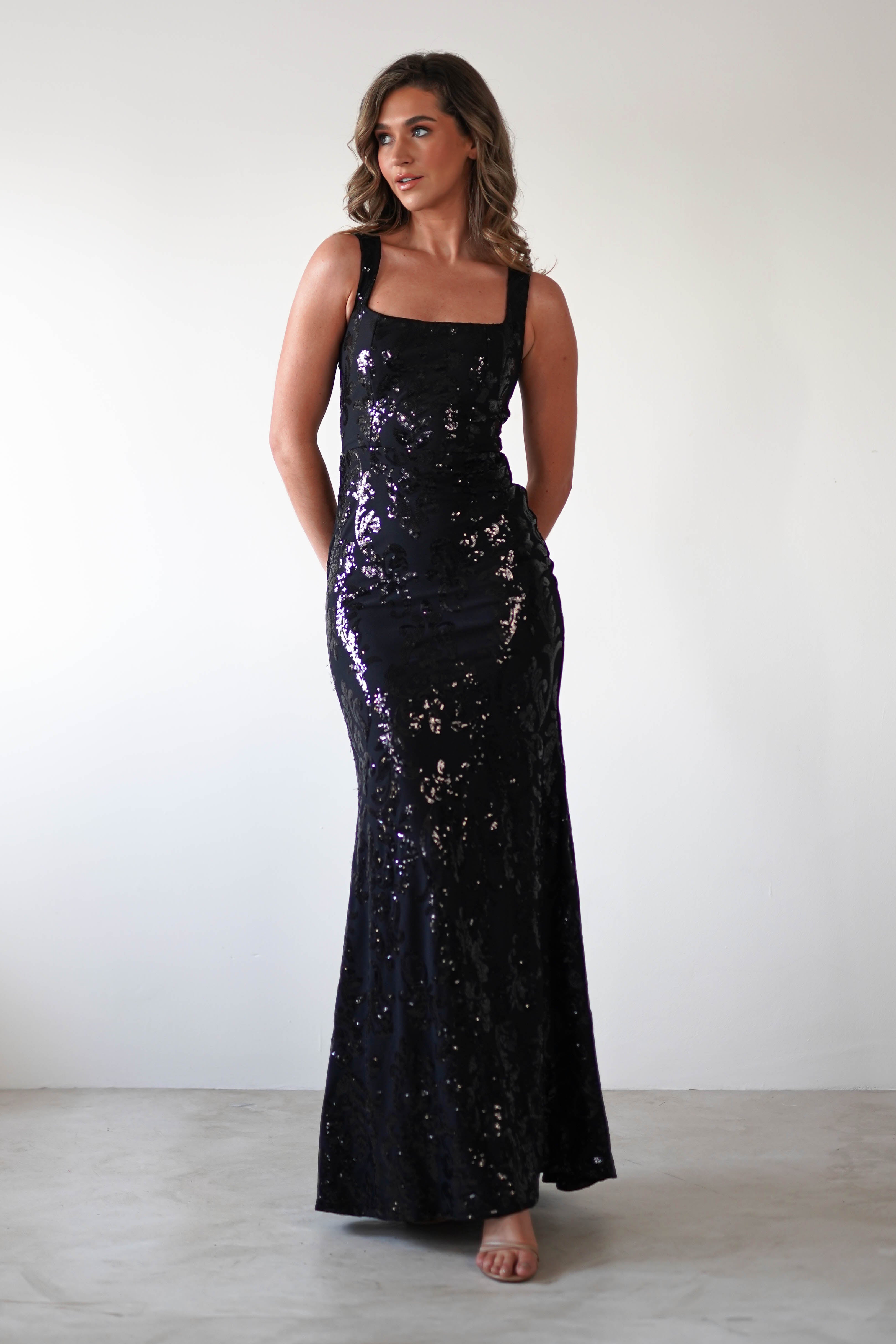 A woman in a black gown with sequin detailing and a square neckline stands against a plain white background. The dress is floor-length with a fitted bodice and wide straps. She has long, wavy hair and poses with her hands behind her back.