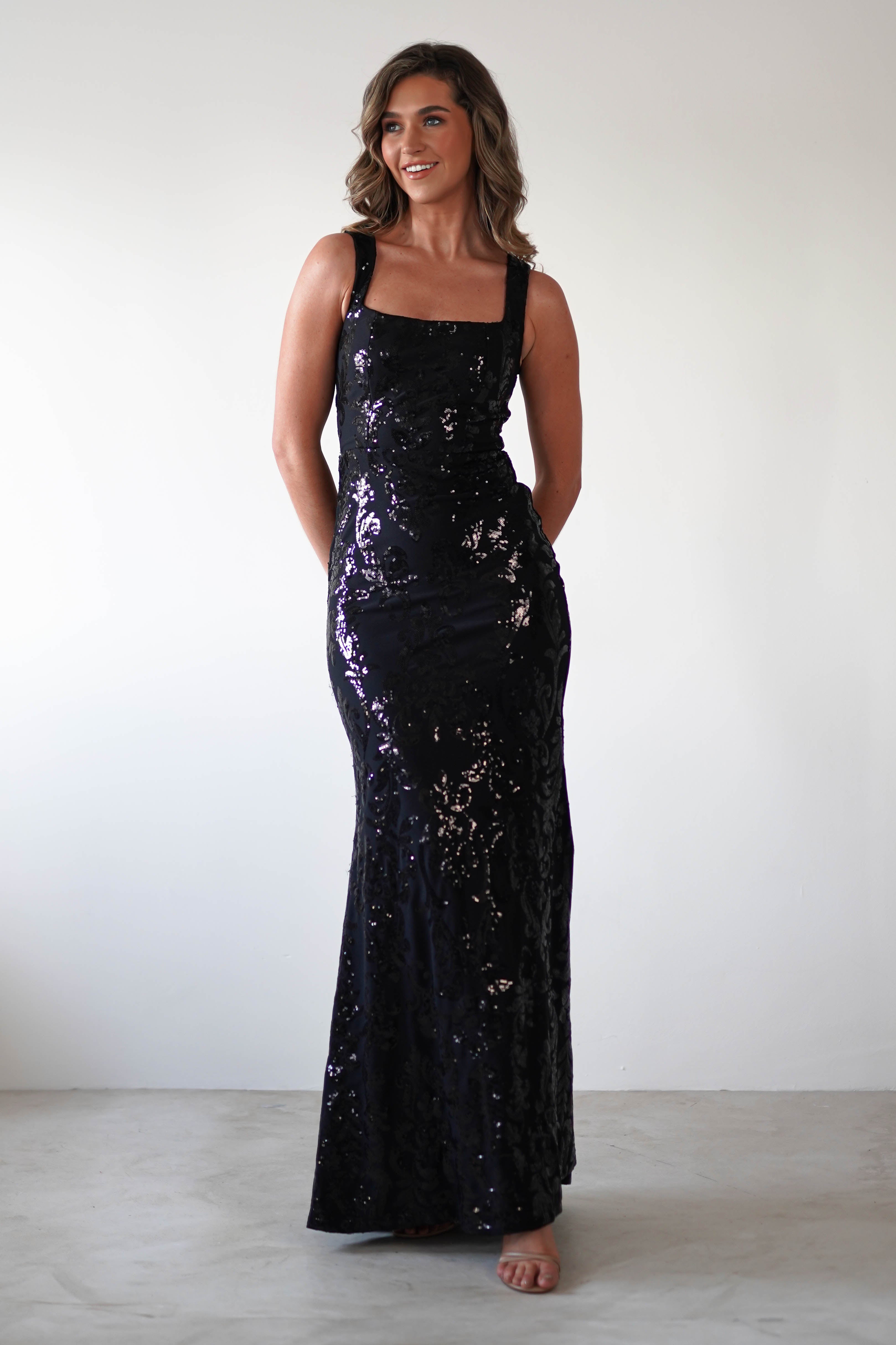 A person wearing a long, sleeveless black gown with a square neckline and shimmering sequin detailing stands against a plain white background. They are looking slightly to the side with a soft smile, hands clasped gently behind.