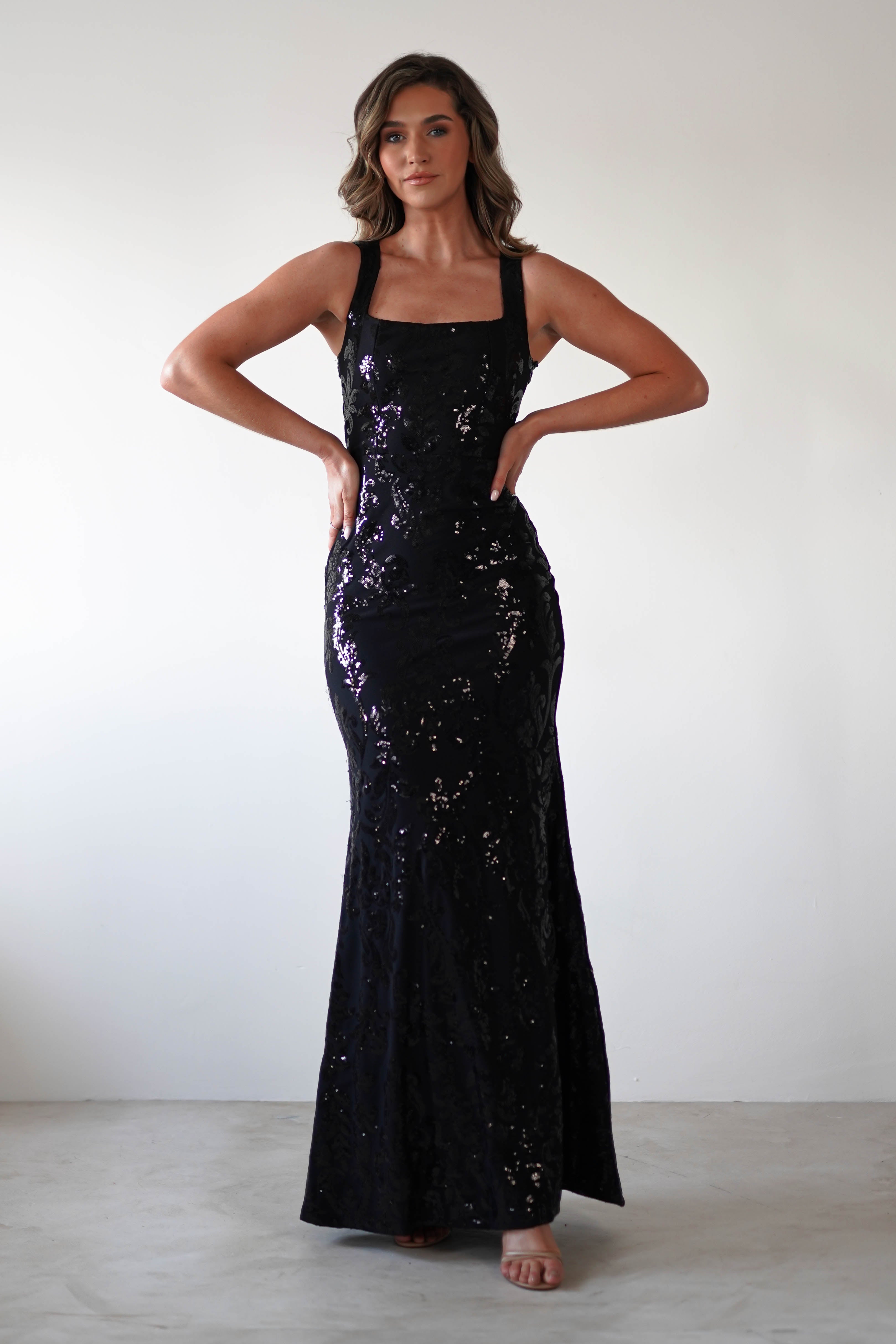A woman in a long, sleeveless black gown with sequin detailing strikes a pose against a plain white background. With wavy brown hair and subtle makeup, she exudes elegance. The dress features a fitted silhouette, square neckline, and slightly flared hem, epitomizing timeless glamour.