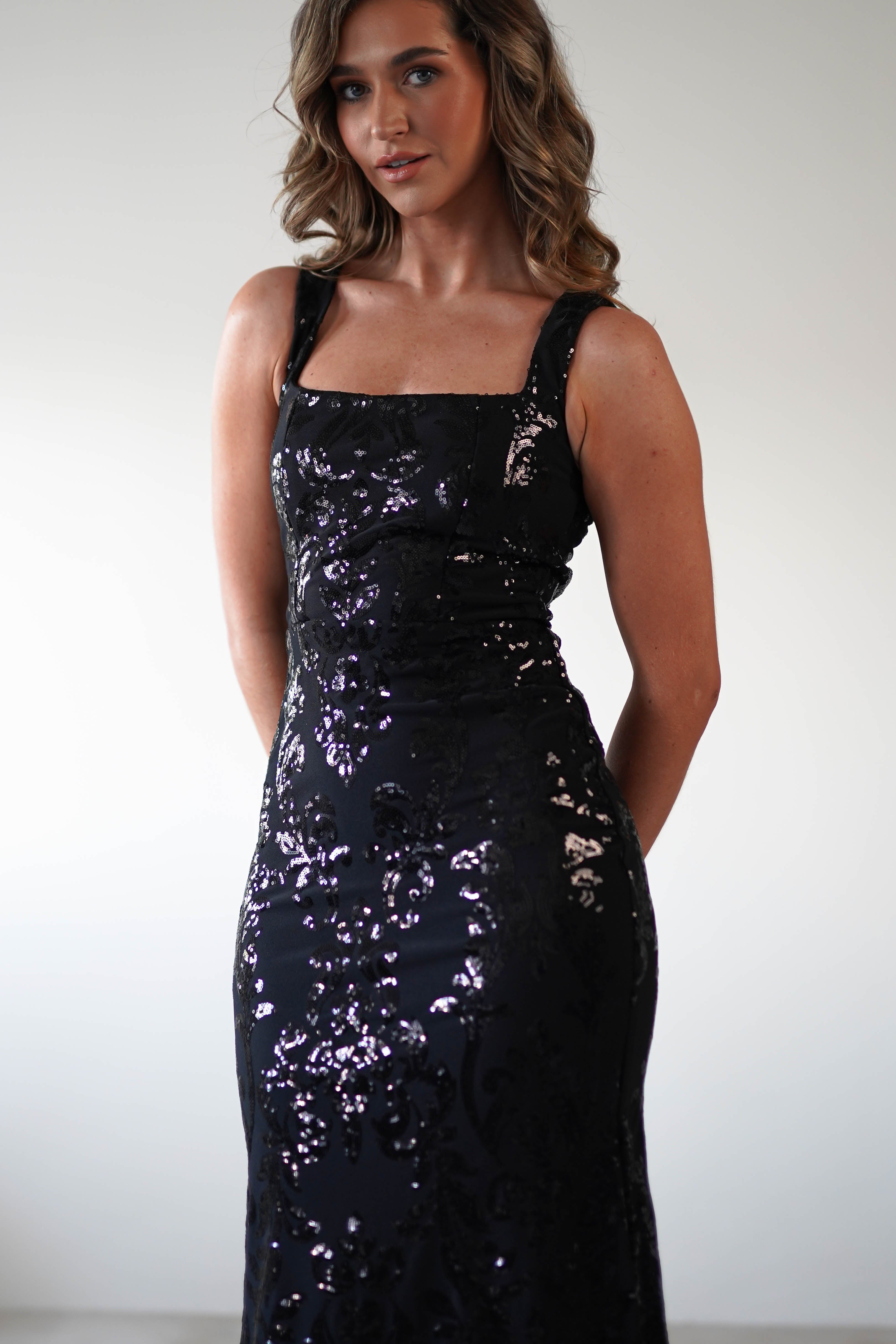 A woman with wavy brown hair wears a shimmering black gown with sequin detailing and a square neckline. She stands against a plain white background, looking confidently at the camera with her arms by her sides.