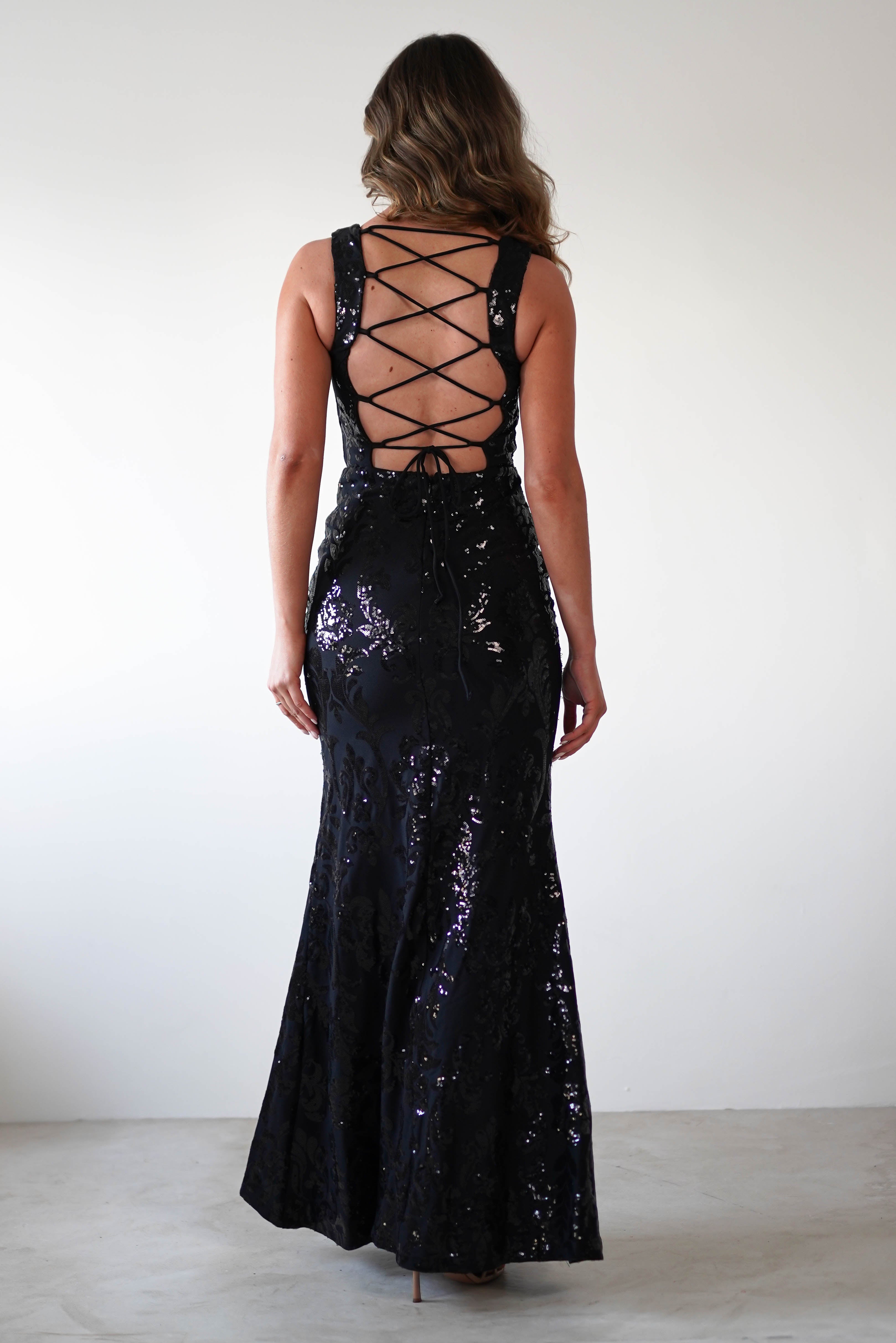 A woman stands against a plain white background, flaunting a floor-length black gown with a square neckline and intricate sequin detailing. Her hair is styled in loose waves, cascading gracefully down her back.