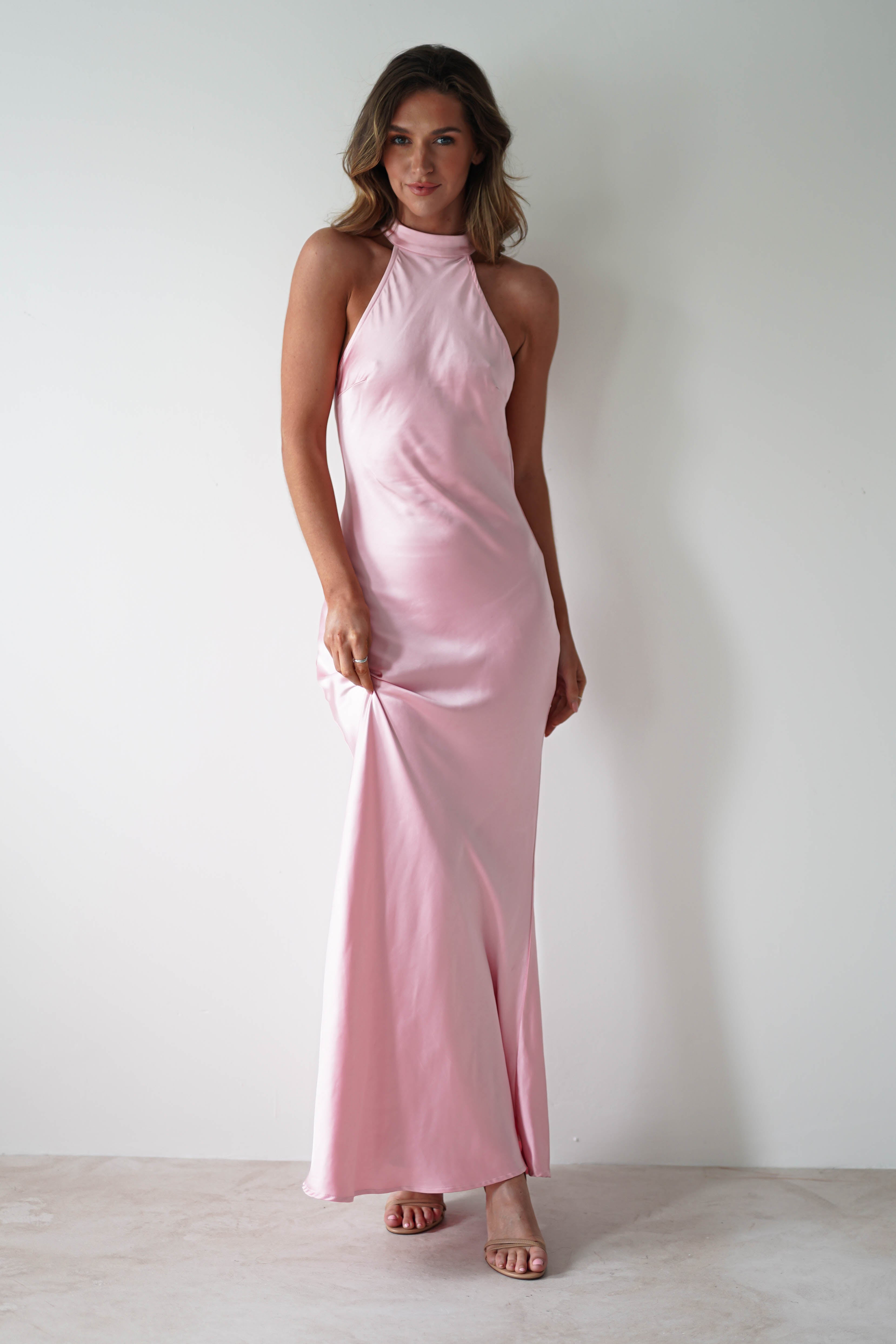 A woman stands against a white wall, wearing a pink satin halter-neck maxi dress with an open-back detail. She holds a portion of the gown with one hand, and her hair falls loosely over her shoulders as she looks directly at the camera.