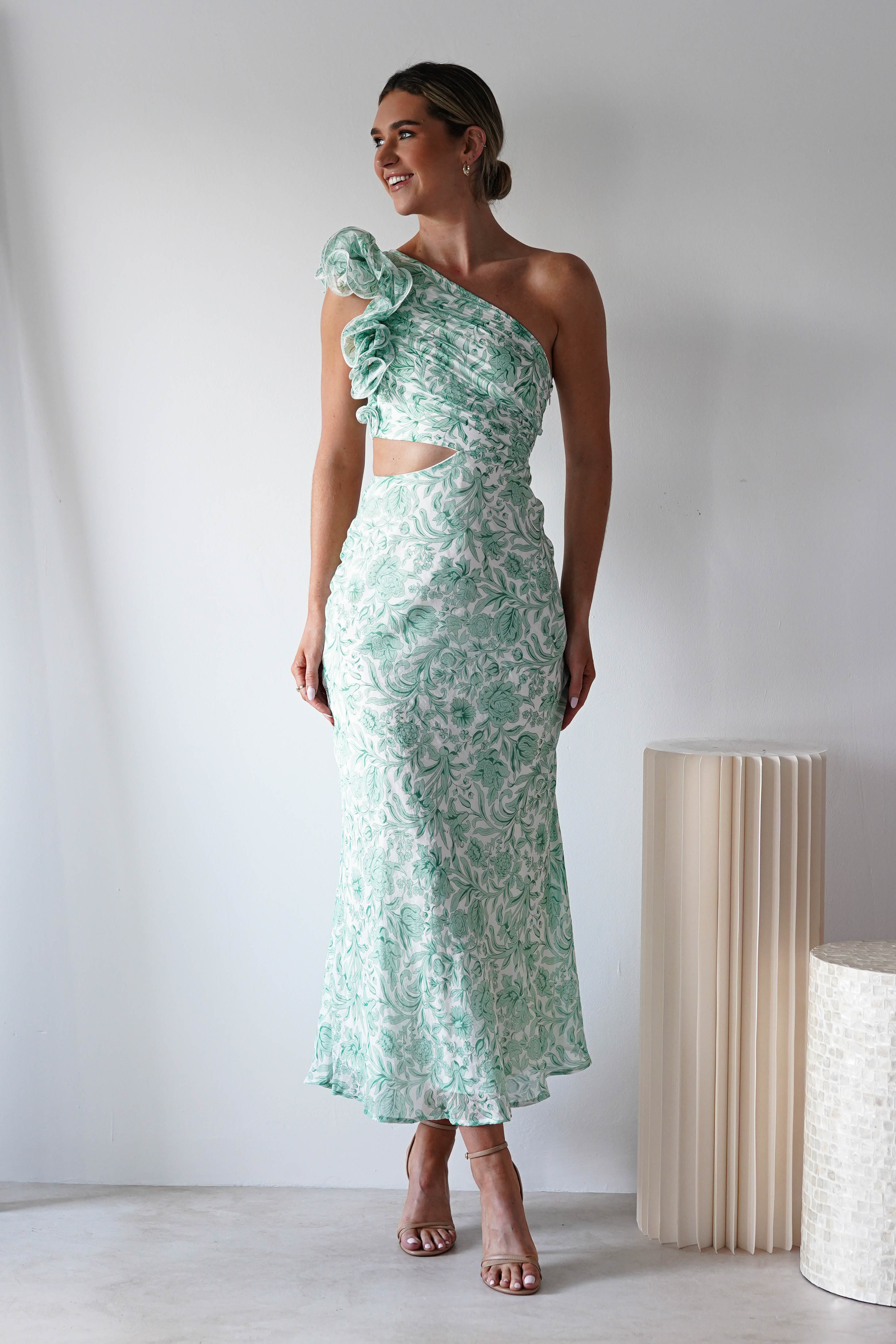 A woman stands in a light-filled room wearing an elegant, one-shoulder dress in mint green floral with textured fabric and a cutout. She has short hair and is smiling, looking to the side. Perfect for special occasions, she stands beside a tall, cylindrical lamp.
