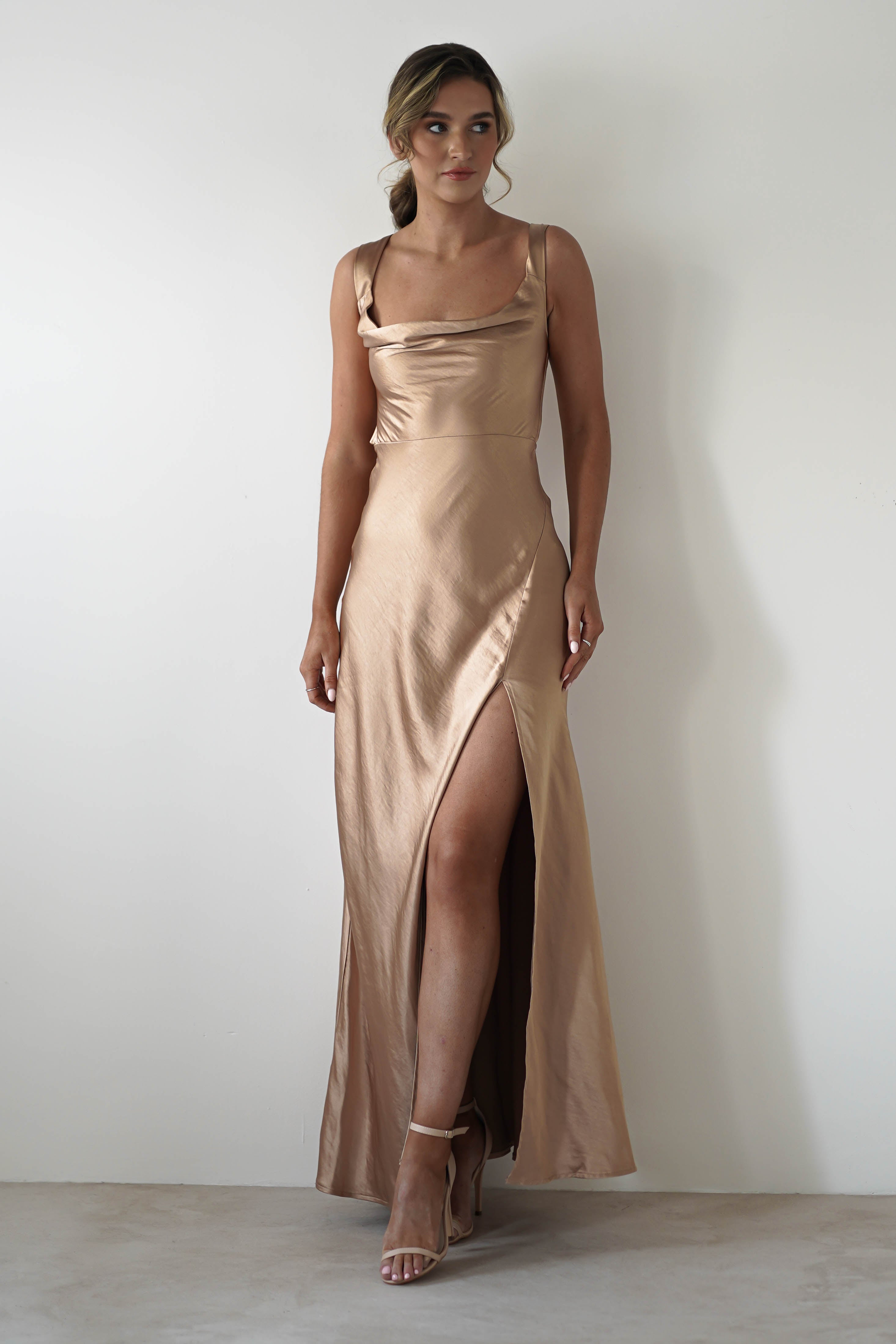 A woman stands against a plain white wall wearing a sleeveless satin fabric gown in champagne, featuring a cowl neckline, high slit, and strappy high-heeled sandals.