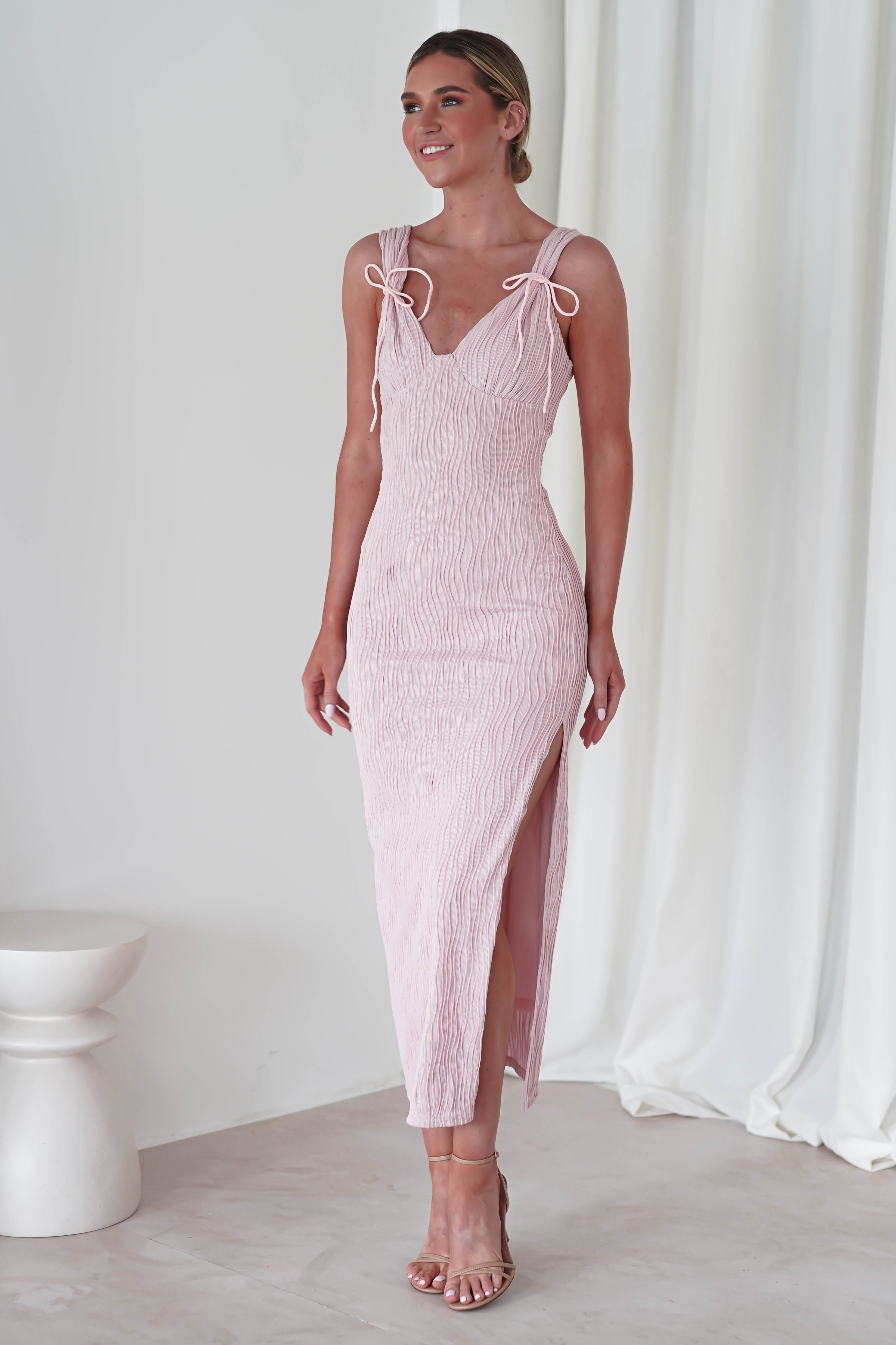 Woman in a light pink midaxi dress with tie straps and a high slit stands against a neutral background. Perfect for special occasions, she is wearing high-heeled sandals and has her hair styled up, embodying the elegance of a wedding guest.