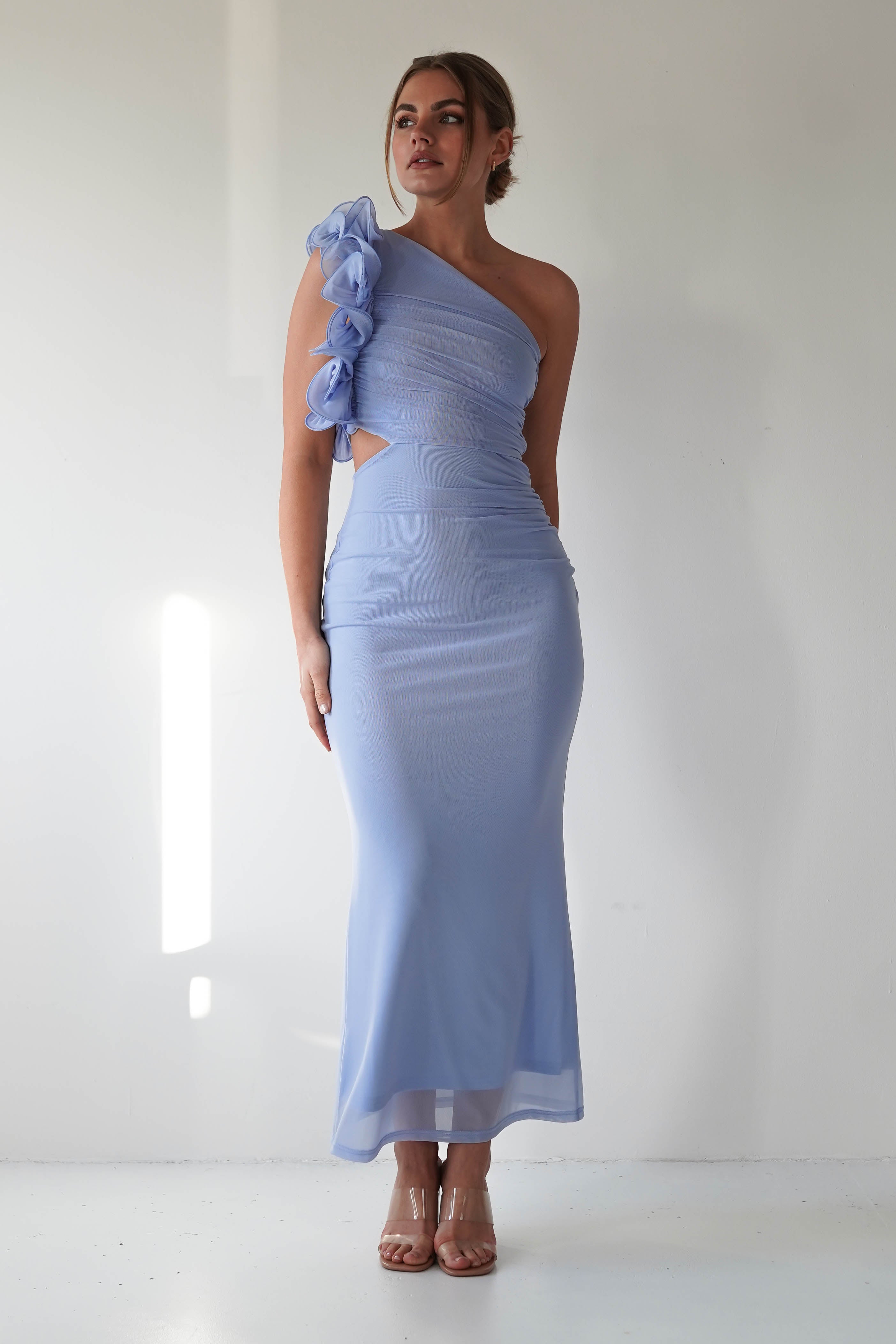 A person wearing a light blue, one-shoulder dress with floral details and a ruffle feature on the strap. The dress, ideal for wedding guest attire, showcases a cutout design flowing to the floor. They stand against a plain white background in high heels.