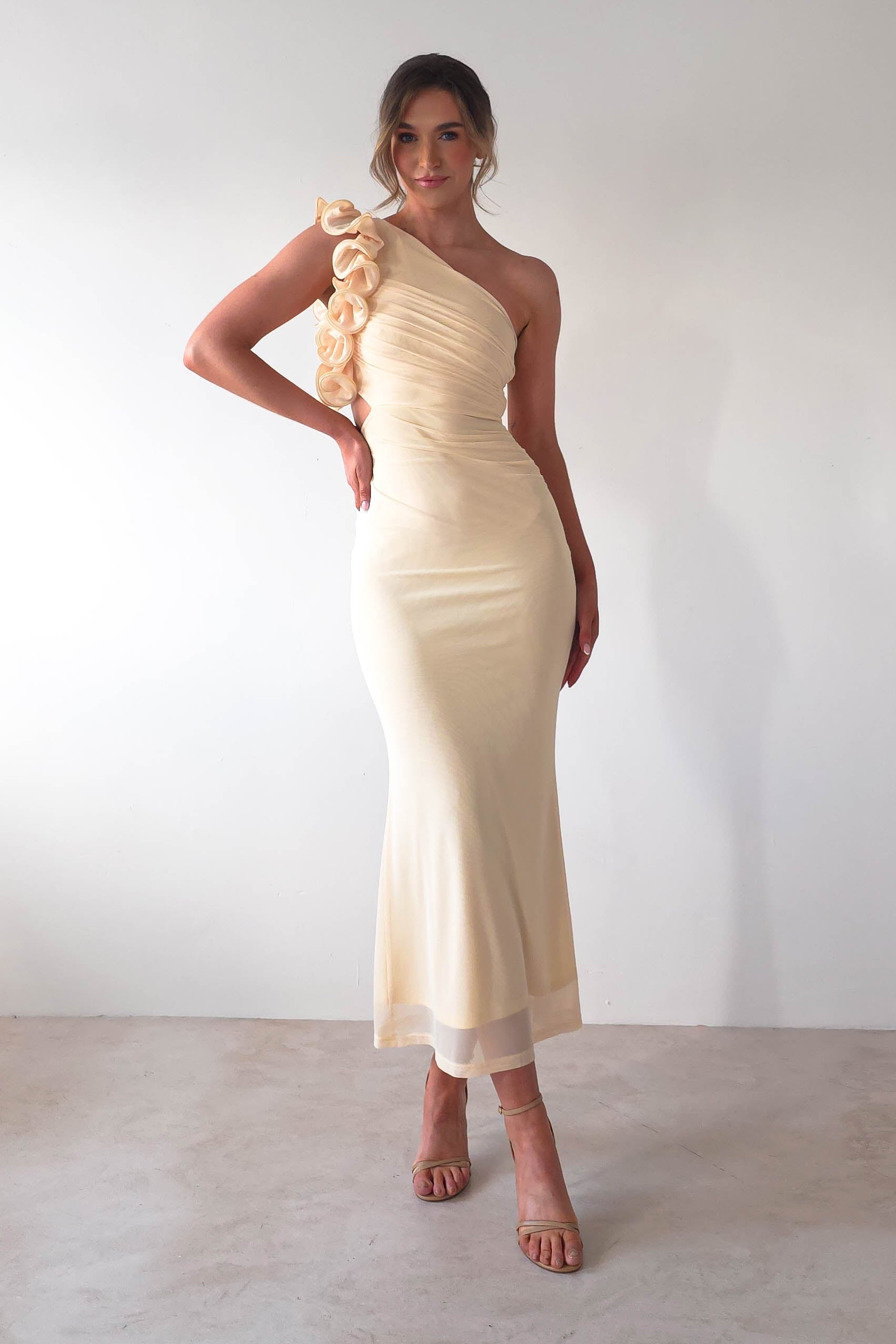 A woman in a fitted, one-shoulder cream midaxi dress with a ruffle feature stands on a plain background. She wears strappy high heels and has styled hair, smiling slightly as she poses with one hand on her hip, perfectly poised as if ready for a wedding guest appearance.