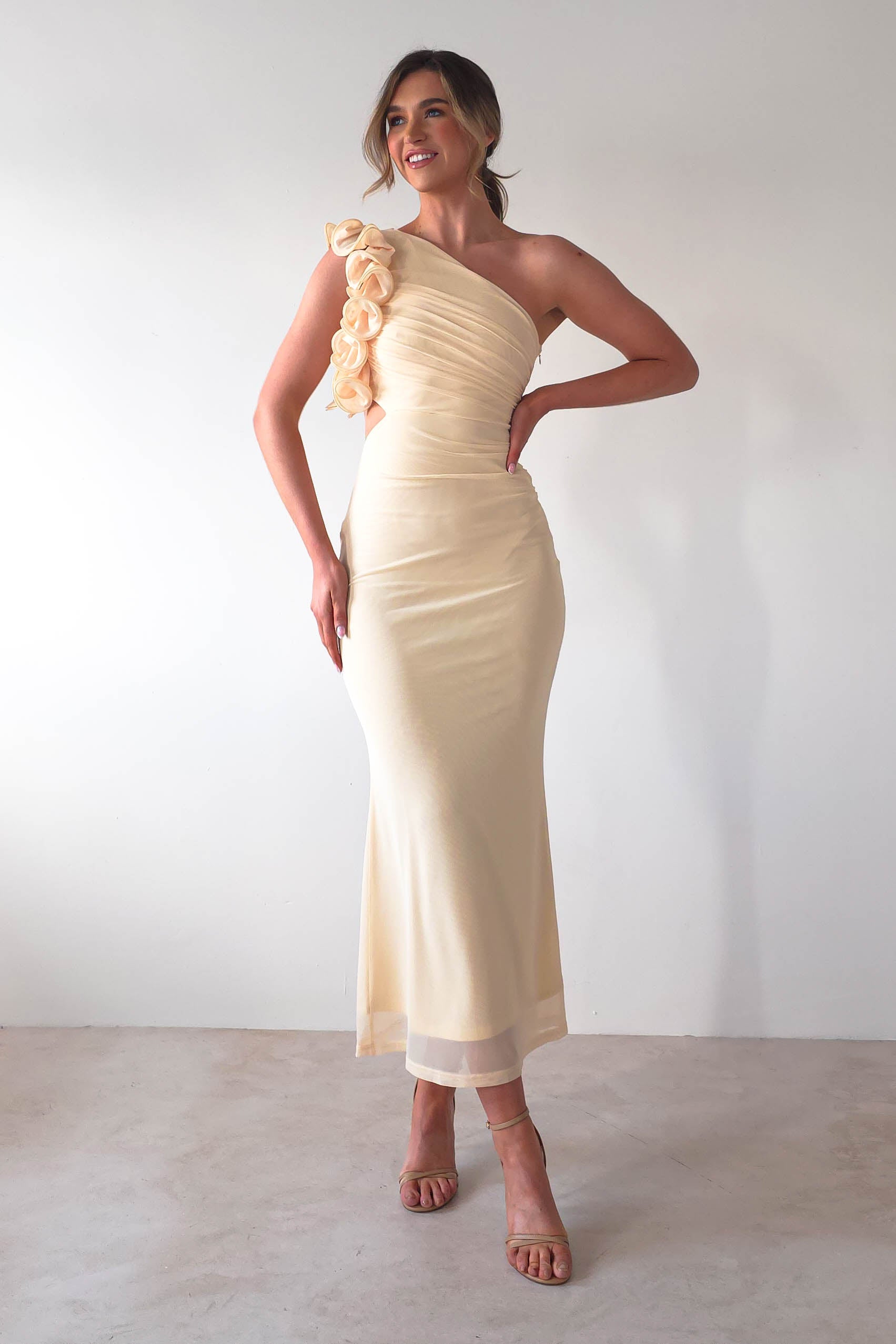 Woman in a form-fitting, one-shoulder beige midaxi dress with floral accents and a subtle ruffle feature on the strap, standing against a plain background. Her hair is styled back, and shes wearing strappy heels, smiling confidently with her hand on her hip – perfect as a wedding guest ensemble.
