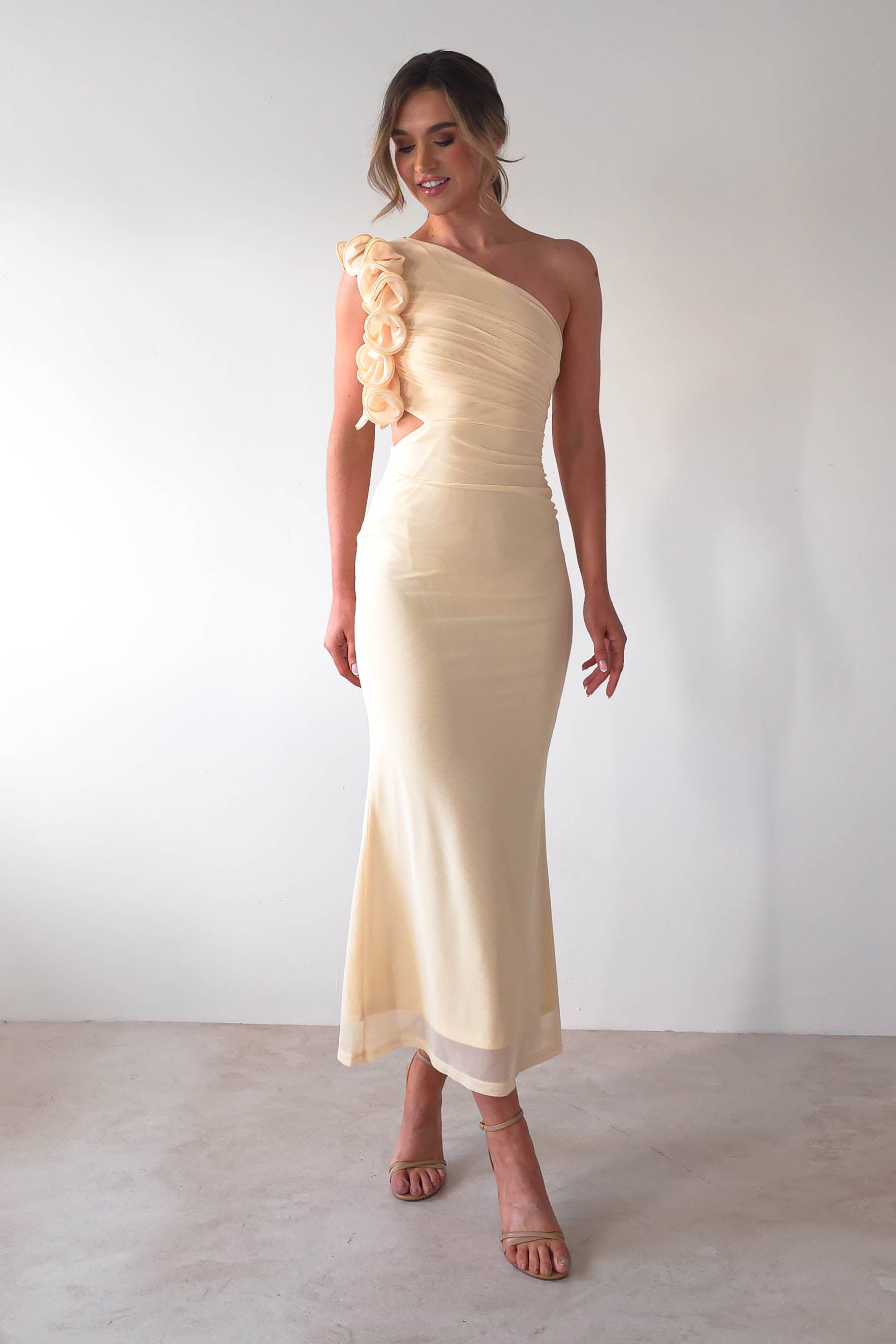 A person wearing an elegant, off-white one-shoulder midaxi dress adorned with floral details along the strap. They have long hair styled up and are standing against a plain background, wearing strappy heels—perfect for a chic wedding guest look.