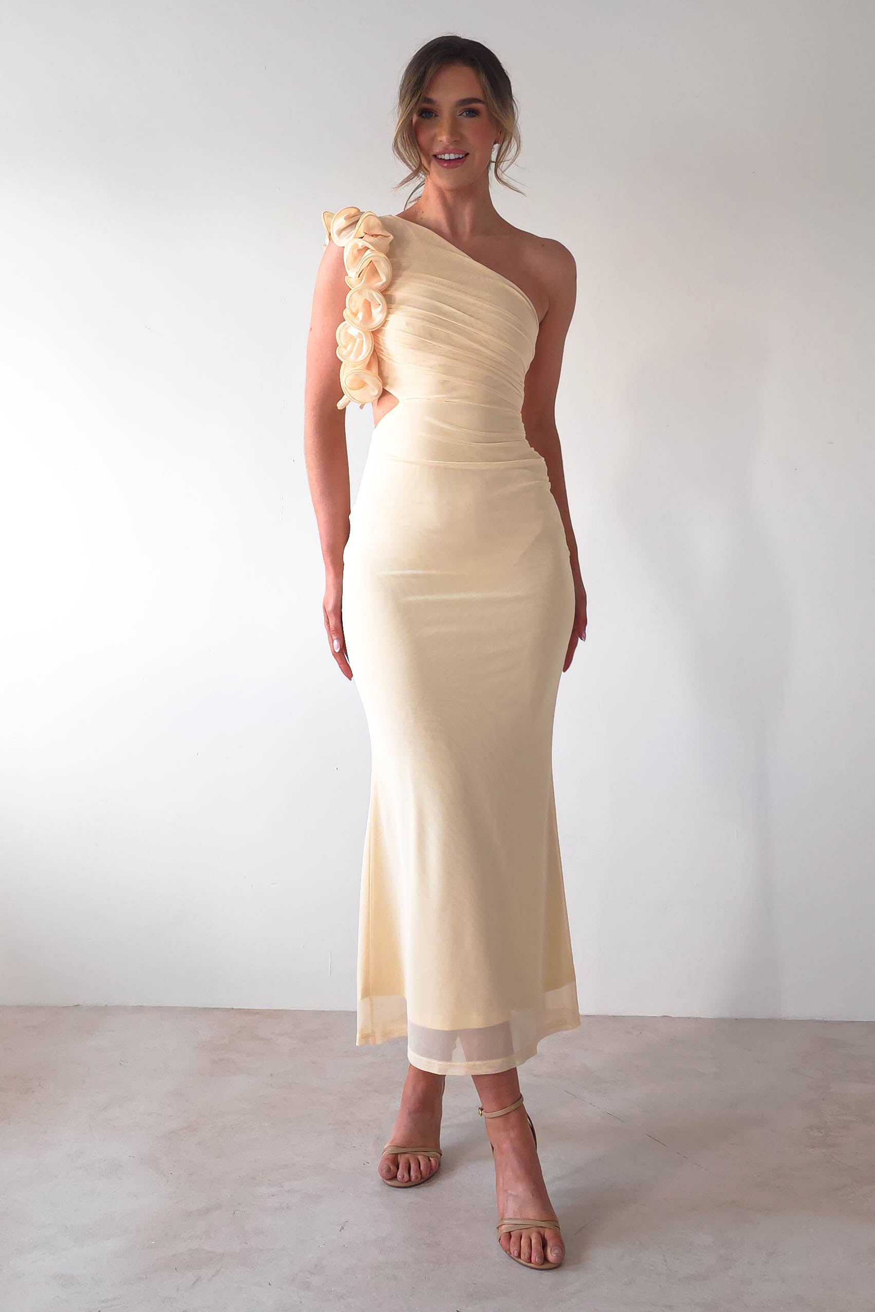 A woman in a pale yellow midaxi dress with floral ruffle features stands against a plain white background. She wears strappy high heels, and her hair is styled up, making her the perfect wedding guest.
