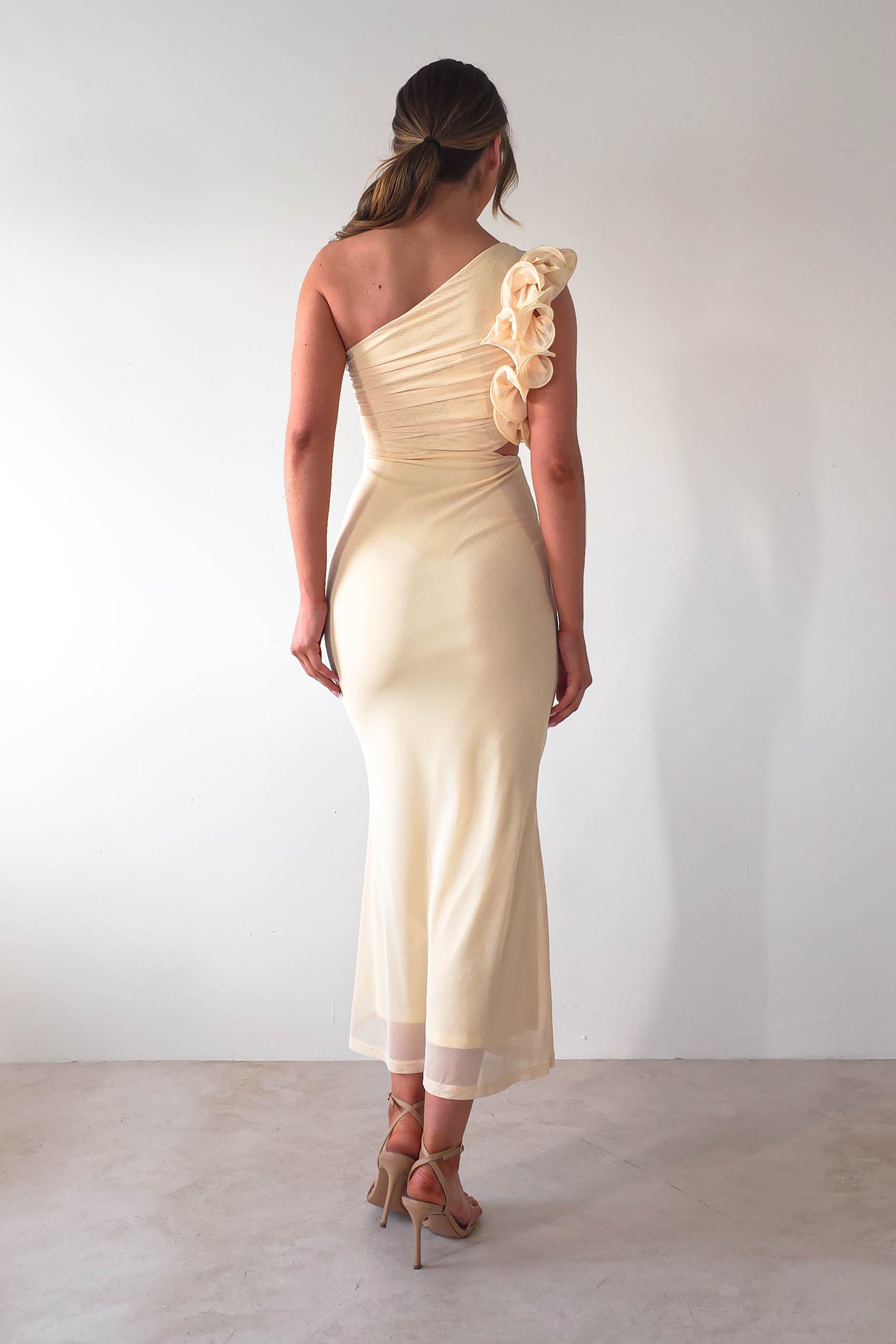 A woman in an elegant, asymmetrical, off-white midaxi dress with floral details on the shoulder stands with her back to the camera. The dress flows gracefully to ankle length. Perfect for a wedding guest, she wears tan high heels and her hair is styled in a low ponytail.