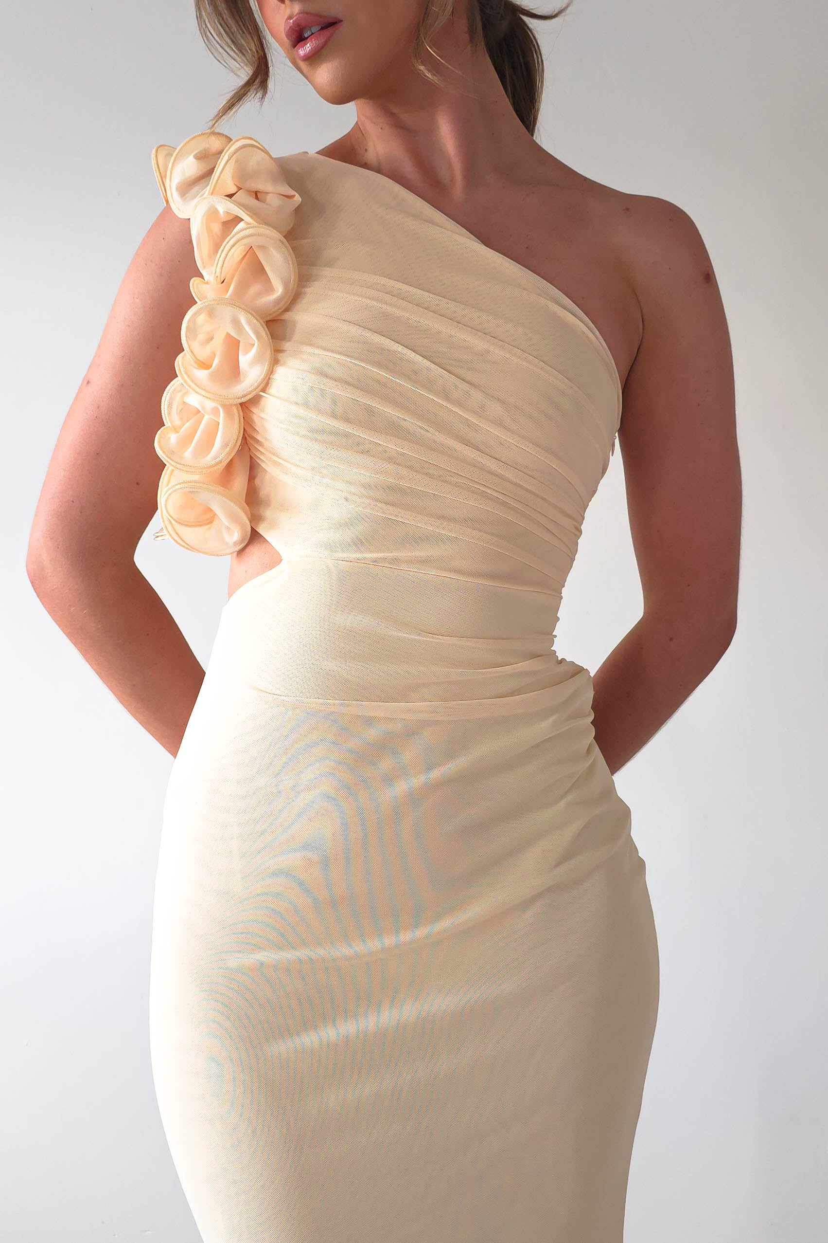 A person poses in an elegant midaxi dress, featuring a one-shoulder design with floral details and a ruffle feature on the strap. The cream-colored dress boasts a fitted style perfect for a wedding guest, exuding sophistication against the plain, light background.