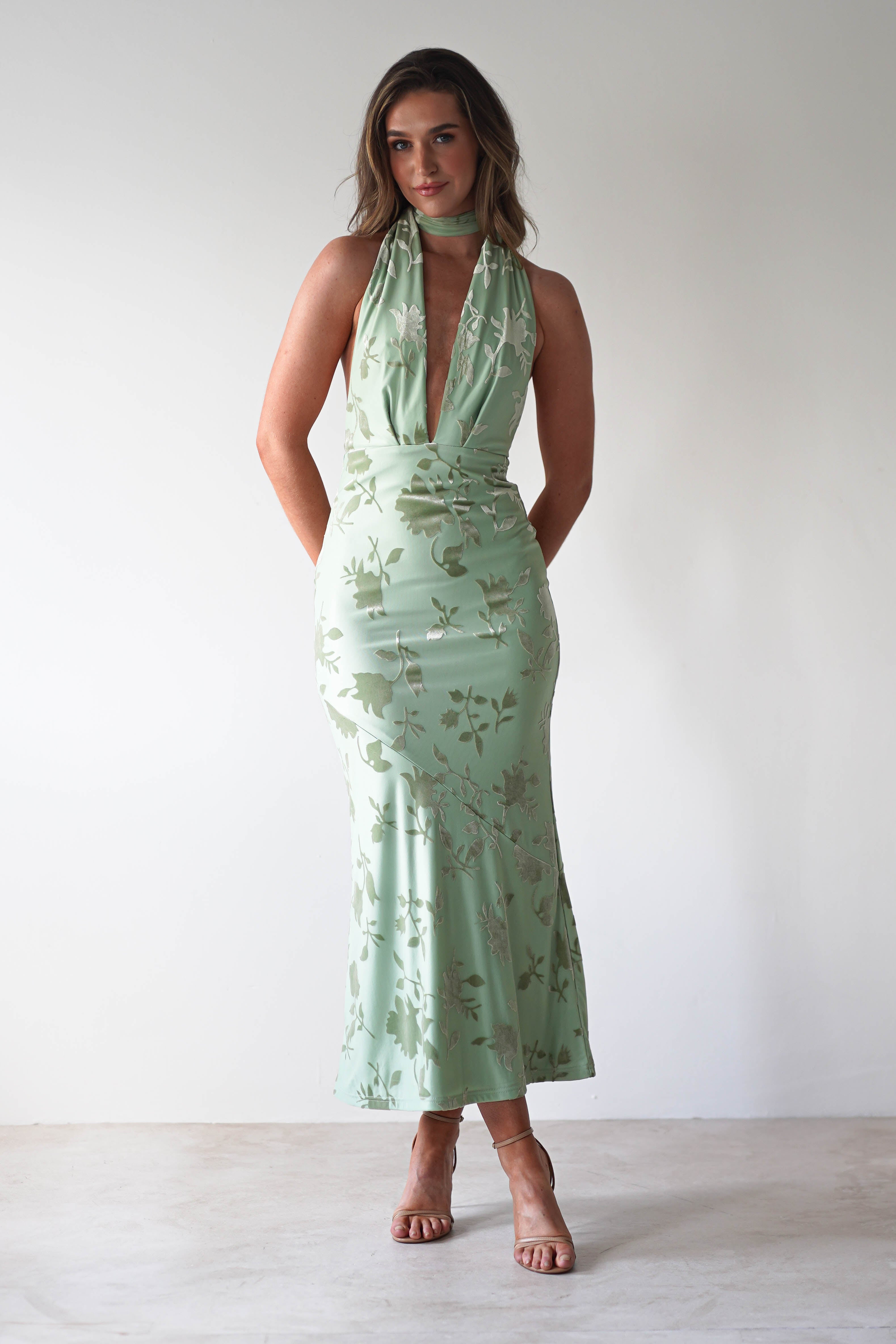 A person poses in a light green sleeveless dress with a deep V-neck and floral pattern, perfect for a wedding guest. The jacquard fabric dress is fitted and ankle-length, featuring adjustable tie straps. They have long, wavy hair and stand against a plain white background.