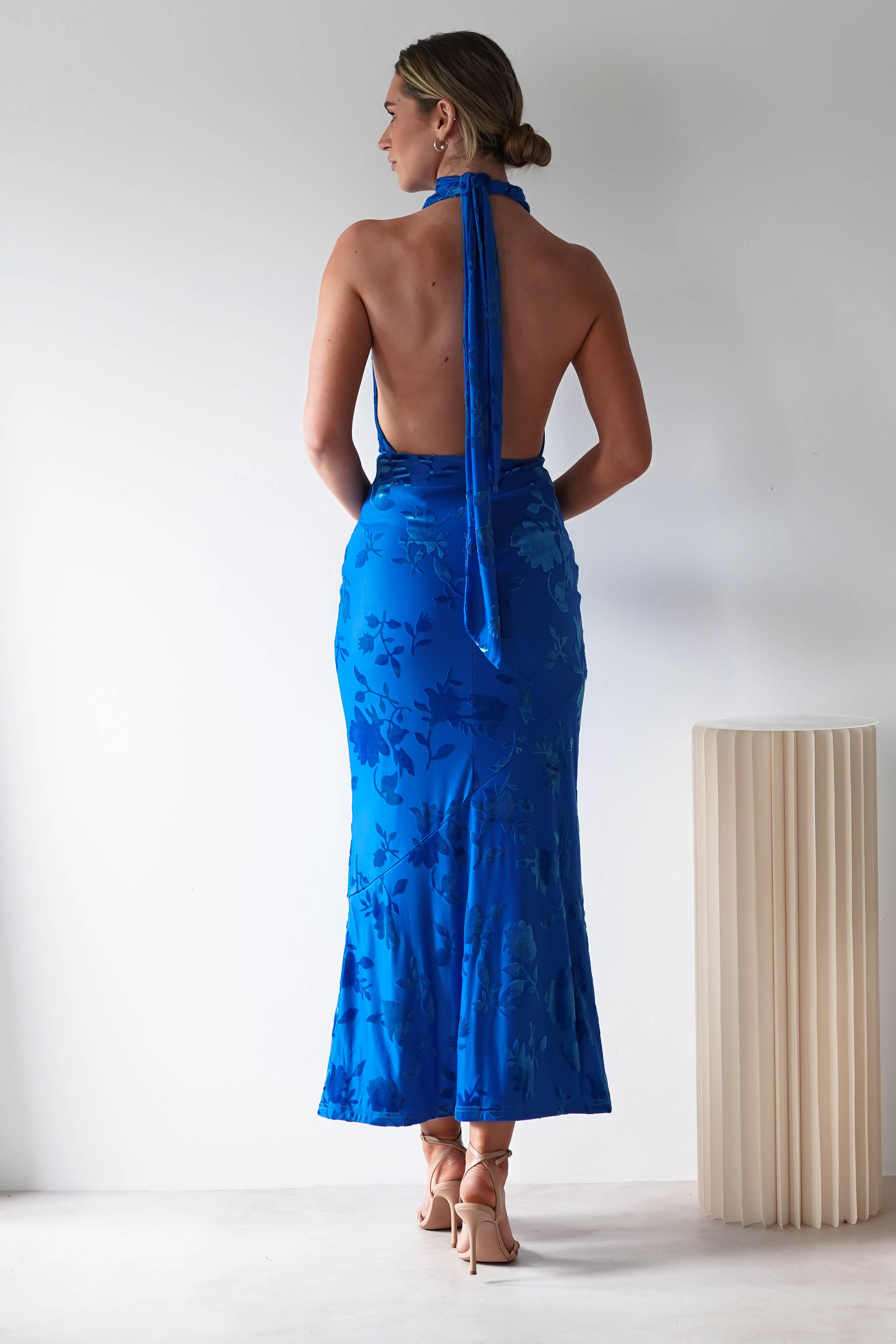 A woman, perfect as a wedding guest, graces a vibrant blue backless halter dress featuring floral patterns made from luxurious jacquard fabric. Standing near a ribbed floor lamp, her hair is styled in a low bun and she complements her look with beige heels against a simple white background.