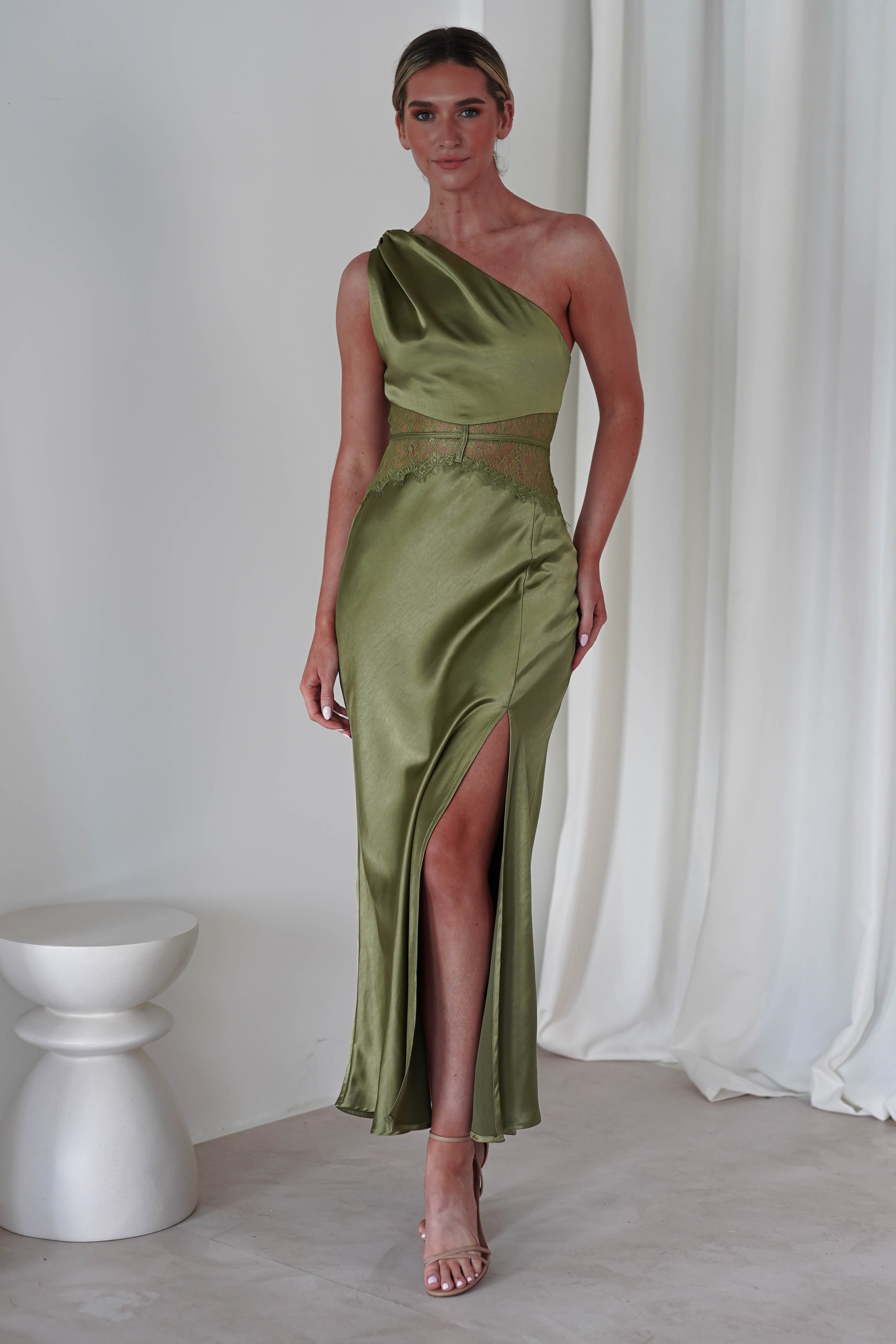A person in an elegant, one-shoulder satin maxi gown featuring a thigh-high slit and delicate lace detailing stands indoors on a light-colored floor. A minimalist white stool and flowing curtains add to the sophisticated wedding guest setting.