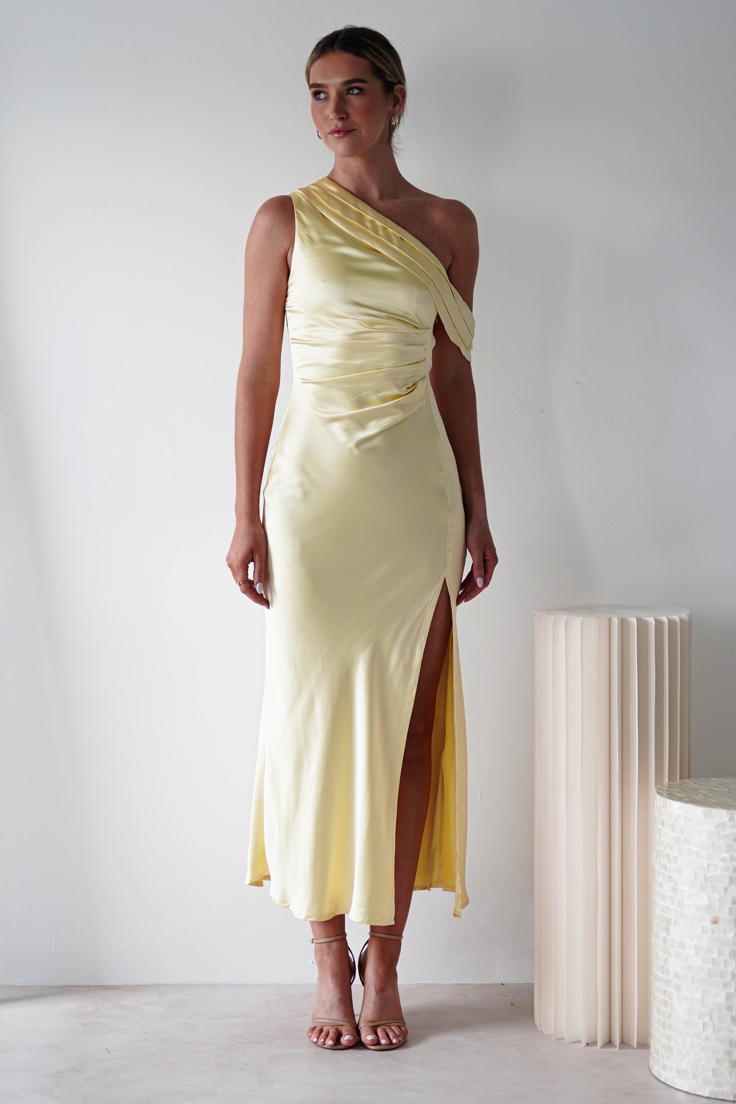 A woman wears a light yellow, one-shoulder satin knit material dress with a side slit, standing indoors against a plain white wall near a textured cylindrical decor piece—perfect for bridesmaids or wedding guests.