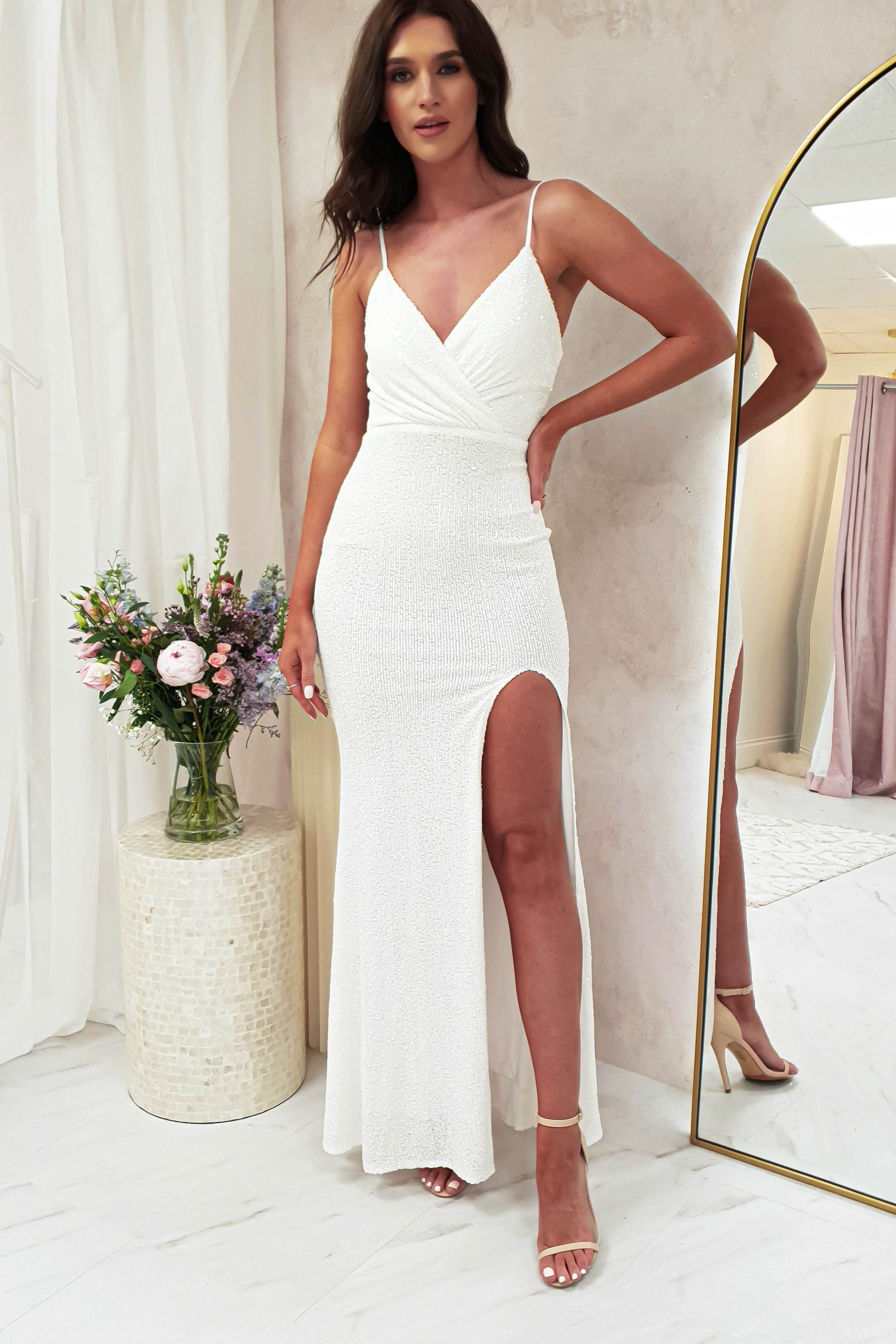 A woman in a luxury fitted, white, floor-length dress with a thigh-high slit stands indoors by a mirror and a vase of flowers, posing with one hand on her hip and wearing strappy heels.