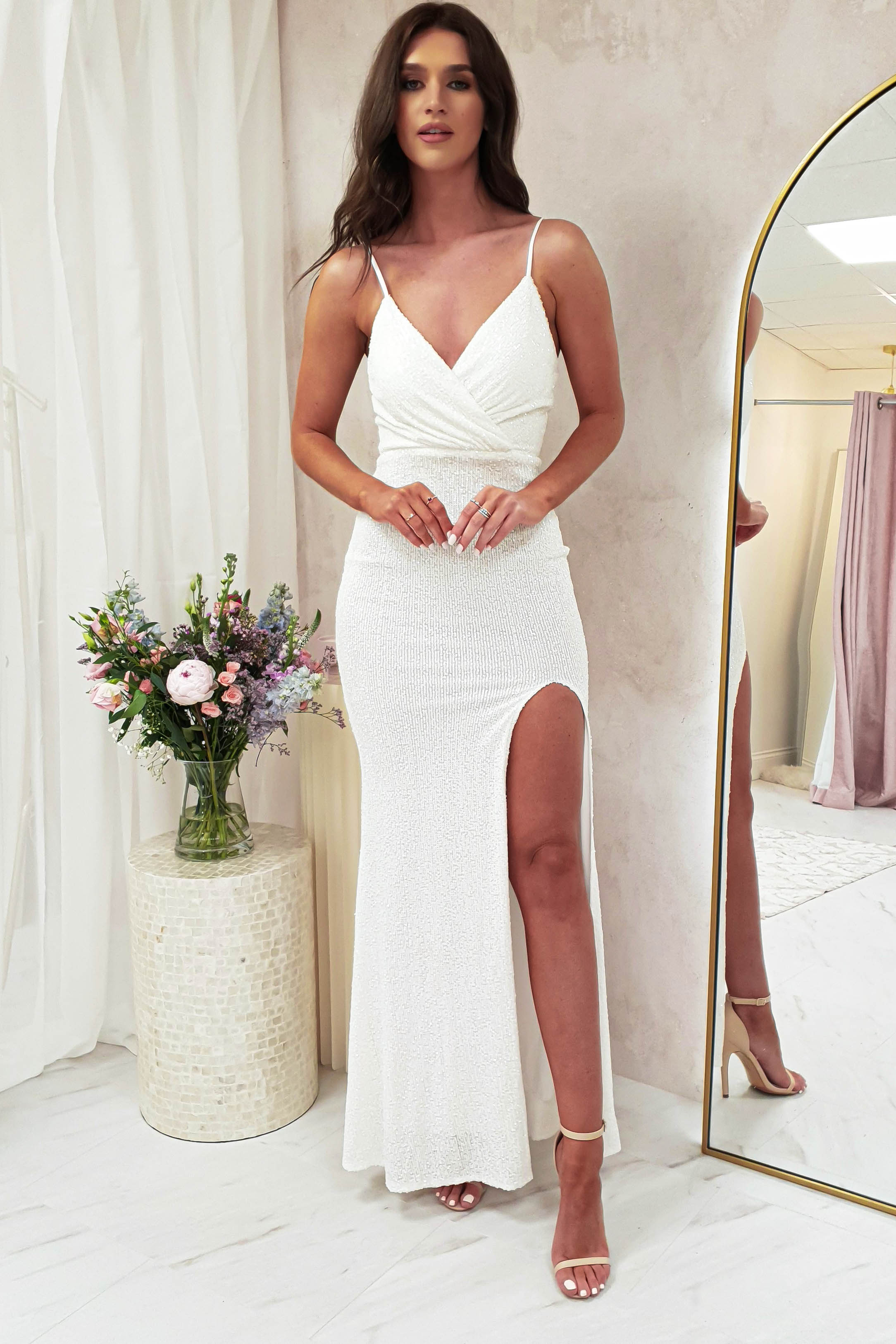 A woman in a luxury fitted white gown with a thigh-high slit and adjustable straps stands indoors beside a round table holding a bouquet of flowers. She wears strappy heels and faces forward, with a large mirror to her right.