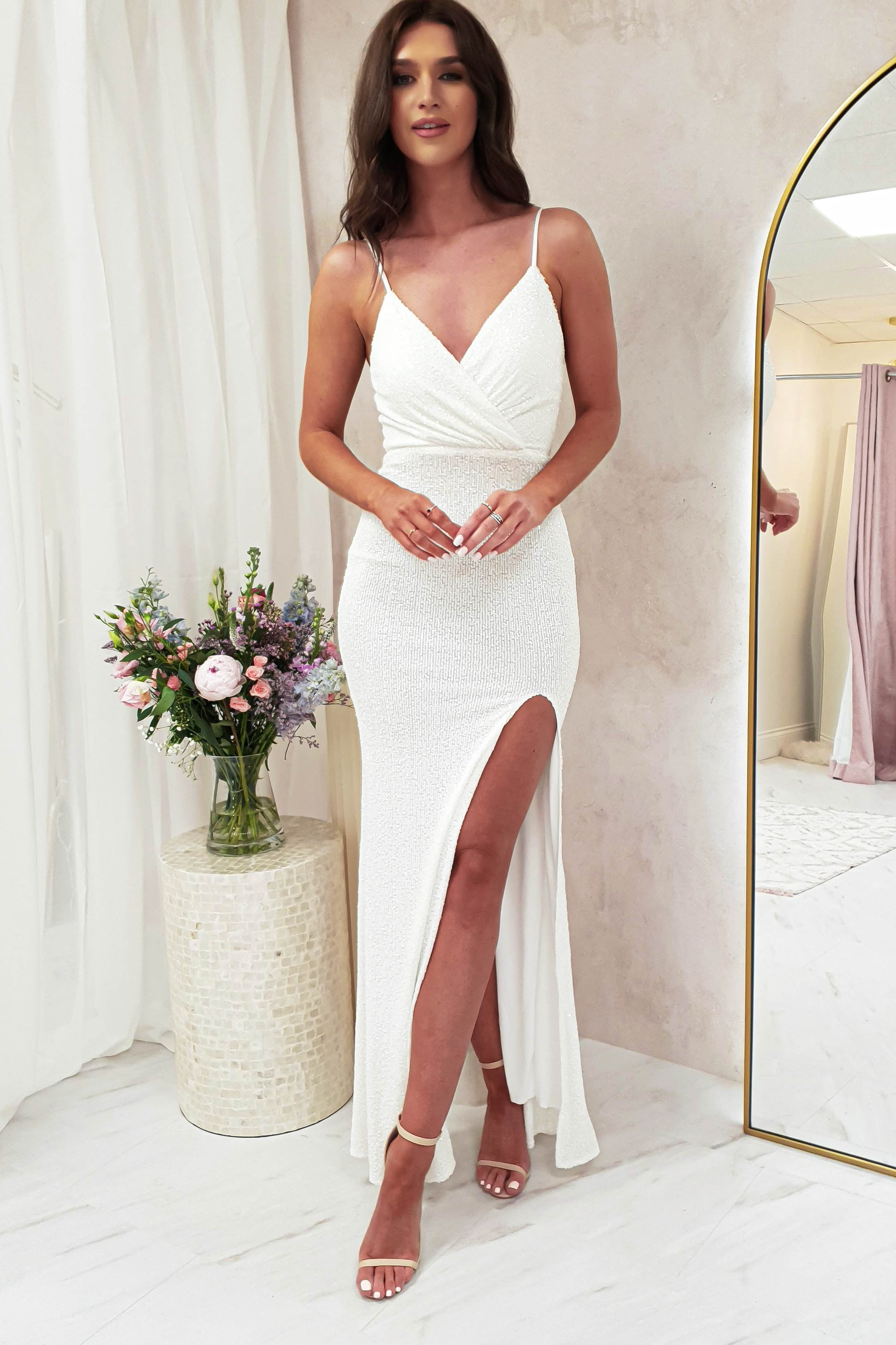 A woman in a luxury fitted white dress with a thigh-high slit and adjustable straps stands indoors near a mirror and a round table with flowers. She wears strappy heels, and her long, wavy brown hair cascades over her shoulders.