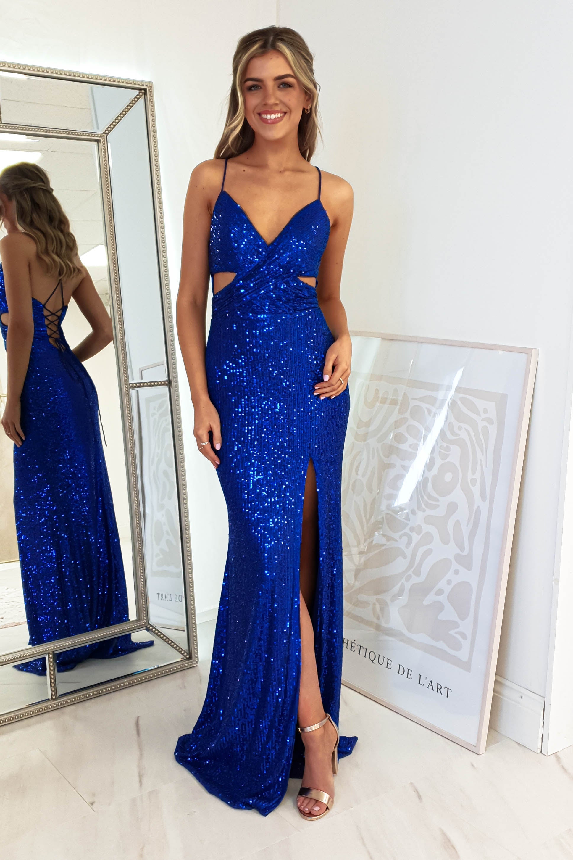 A woman poses in a royal blue sequin gown with cut-outs, a plunging neckline, and a thigh-high slit, standing before a large mirror. She smiles with one hand on her hip. Behind her, framed artwork rests on the floor. She completes the look with strappy heels.