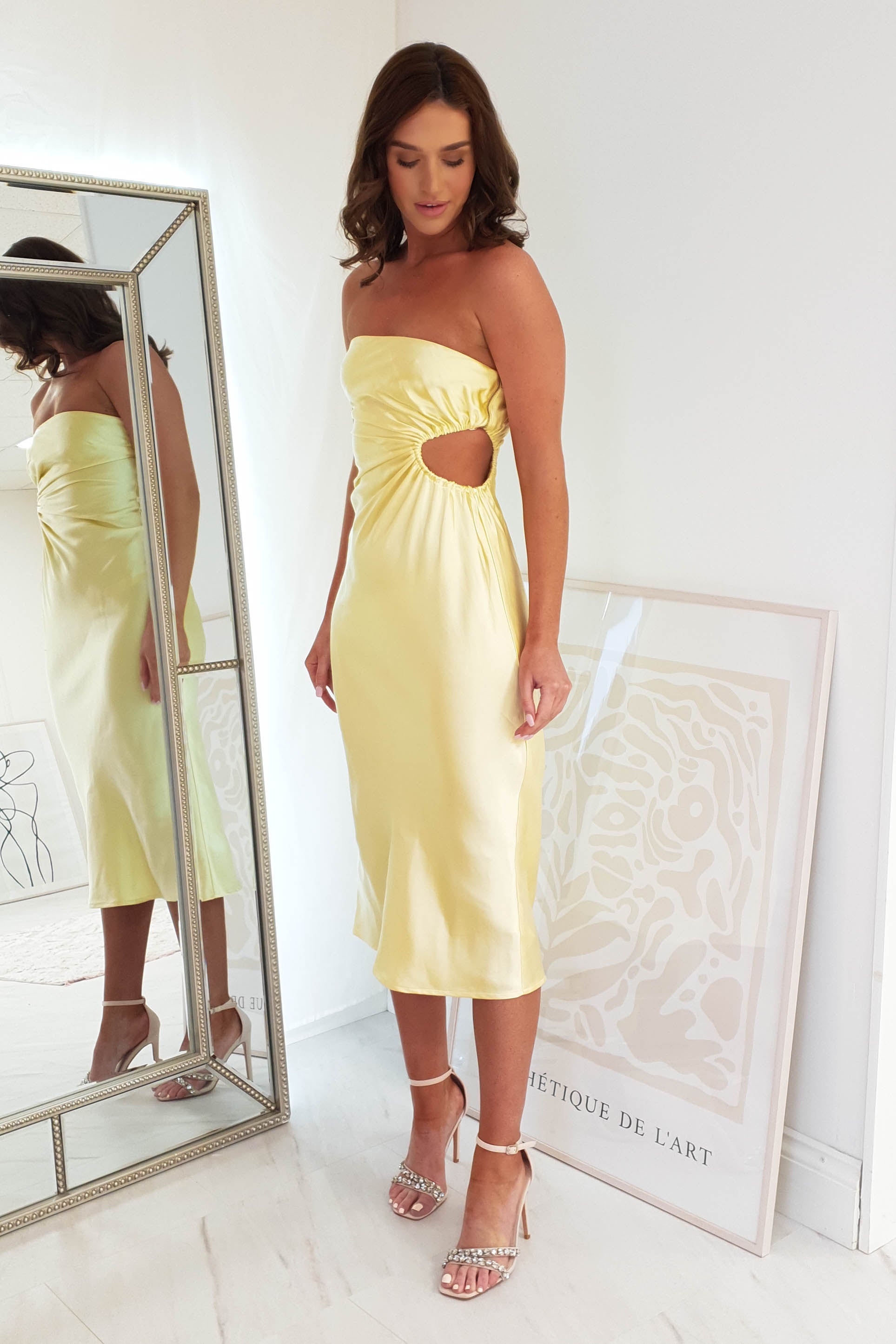 A woman in a satin, yellow strapless midi dress with a side cutout stands in a minimalistic room. Shes admiring her reflection in a large mirror. The space features light-colored walls and flooring, complemented by a framed abstract artwork leaning on the floor.