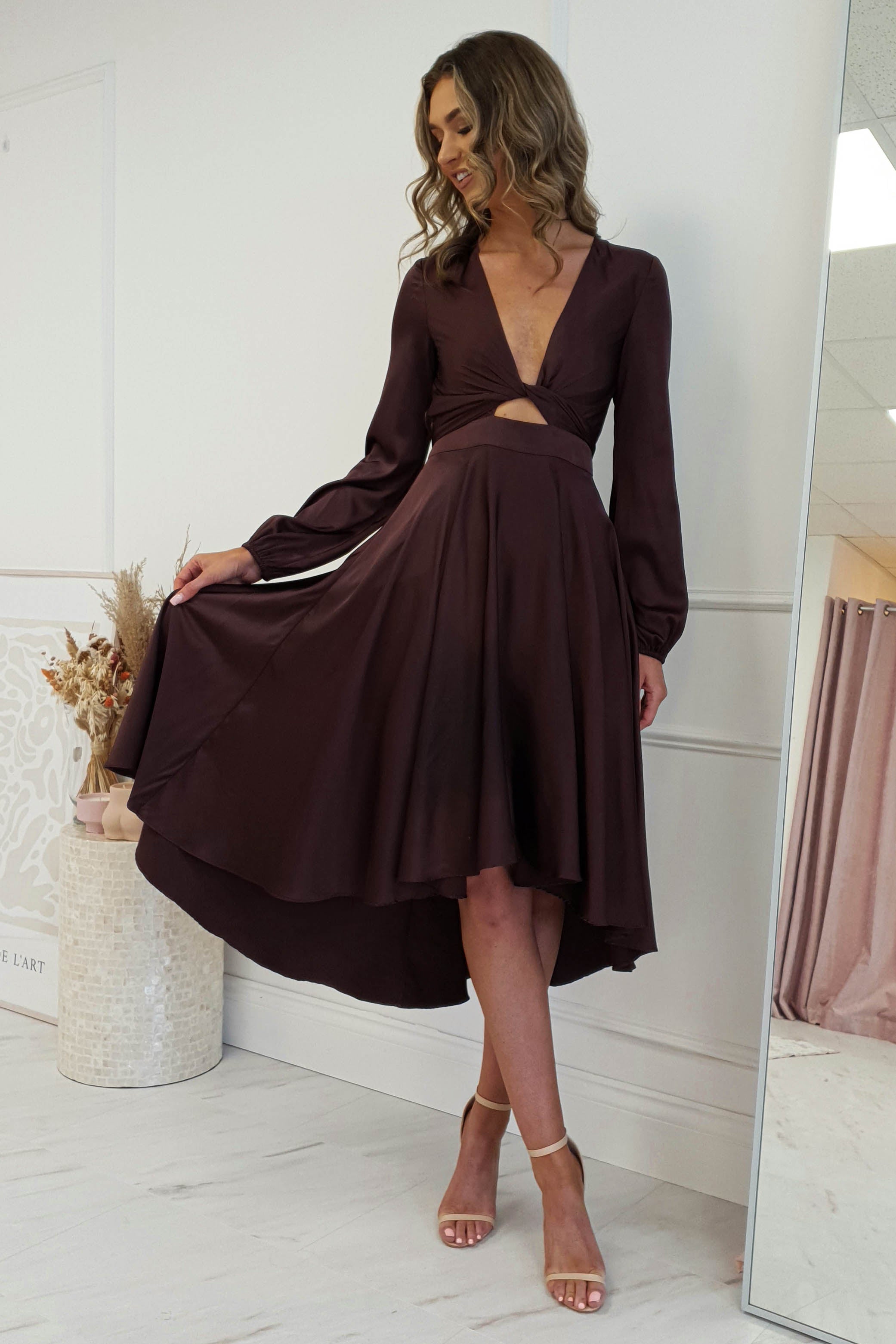 A woman stands indoors, wearing a stylish, long-sleeve midi dress in brown with a high-low hem and cut-out detail. The V-neck design complements her nude heels as she gracefully holds part of the skirt. The rooms neutral tones and decorative elements enhance the elegant scene.