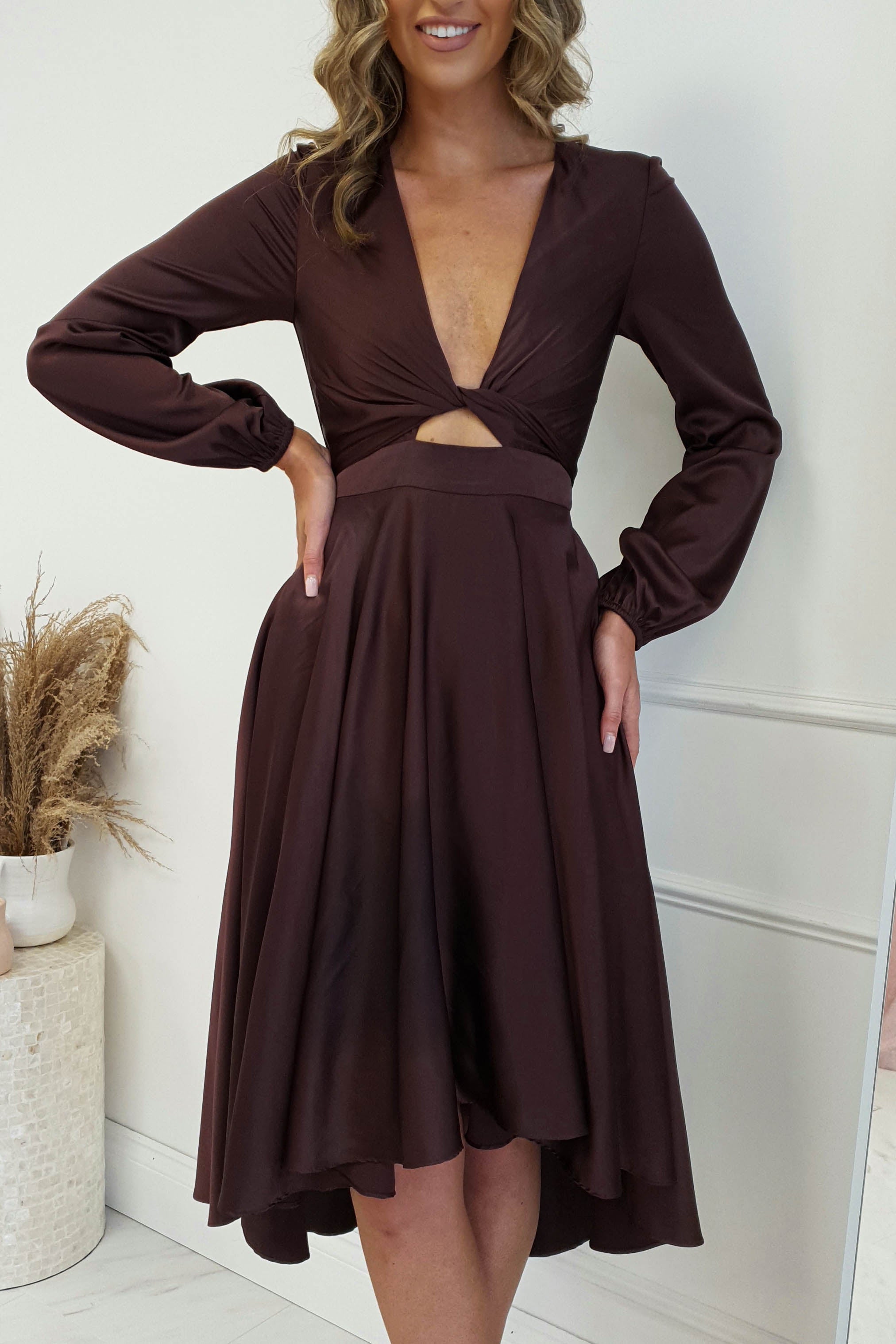 A person stands elegantly in a light room wearing a stylish, long-sleeve midi dress with a deep V-neck and cutout at the waist. The skirt flares gracefully as they pose with one hand on their hip. In the background, a white vase holds dried pampas grass, adding to the chic ambiance.