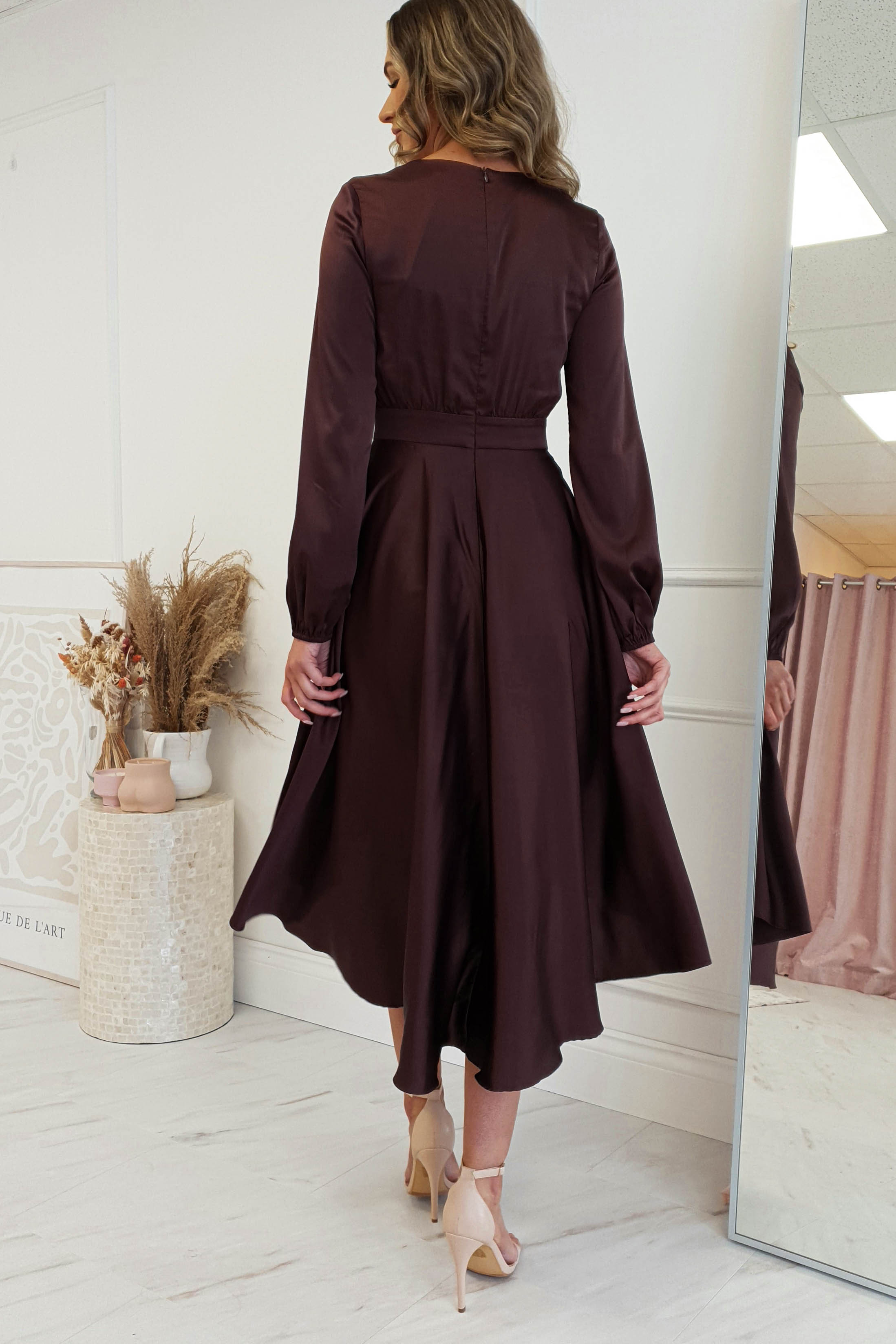 A woman in a stylish, long-sleeve midi dress with a V-neck stands in a room with light walls. The dark brown dress features a fitted waist and flared skirt. She gazes to the side, while a vase with dried plants rests on a nearby decorative stand.
